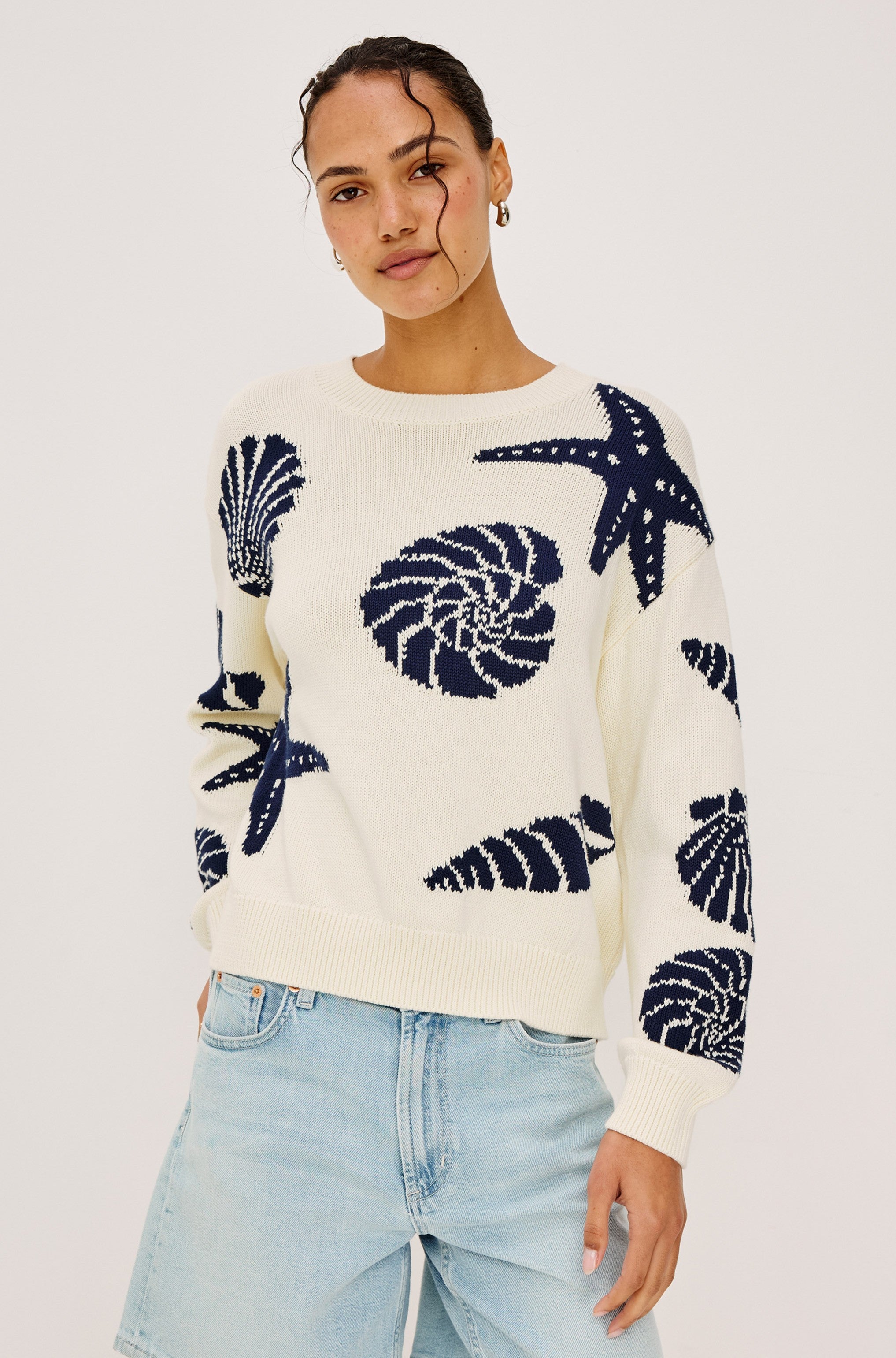 A person models the Rails Zoey Shell Sweater, a white cotton crewneck with navy seashell and starfish patterns, standing against a plain background with hands in light blue jeans pockets and looking at the camera.