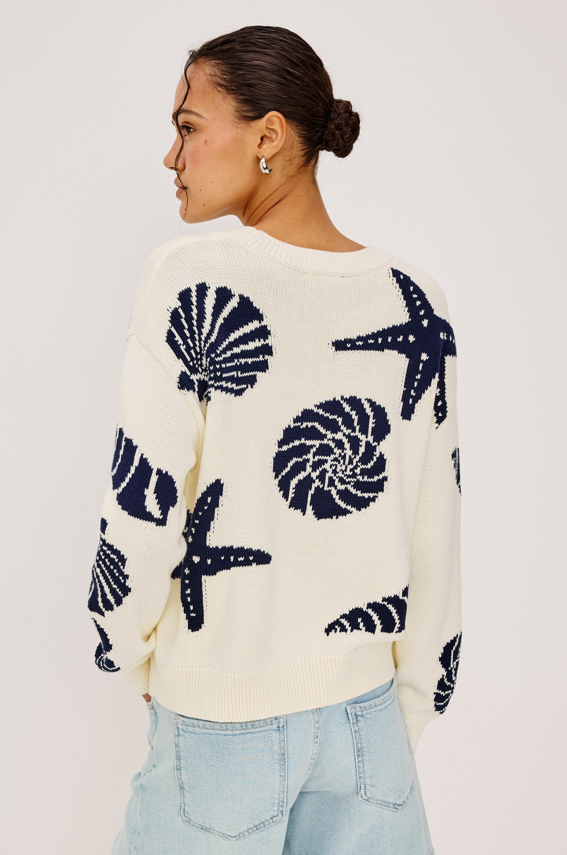 A woman with dark hair in a bun models the Rails Zoey Shell Sweater, a cream crewneck embroidered with navy starfish and shell designs, paired with light blue jeans, standing slightly turned against a plain background.