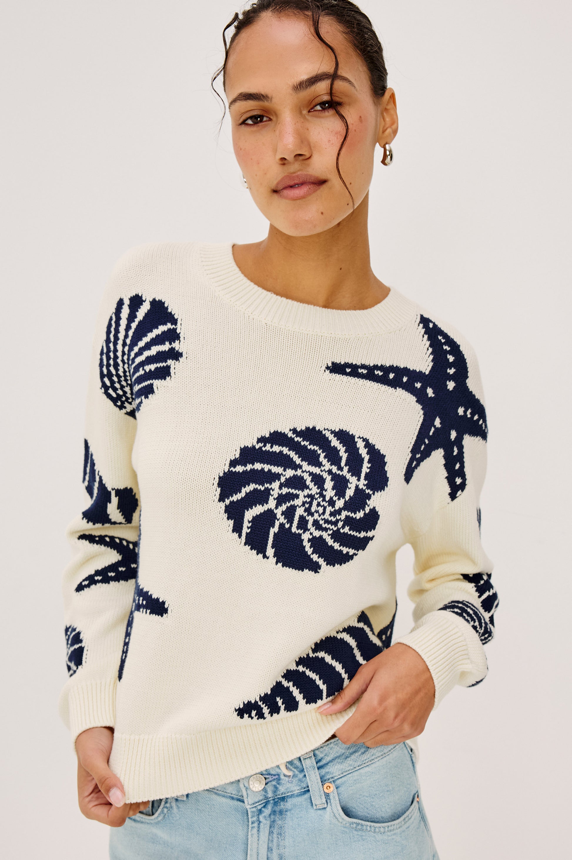 A woman with curly hair in small hoops wears the Rails Zoey Shell Sweater—a cream cotton crewneck featuring navy seashell and starfish motifs—paired with light blue jeans, standing against a plain light background.