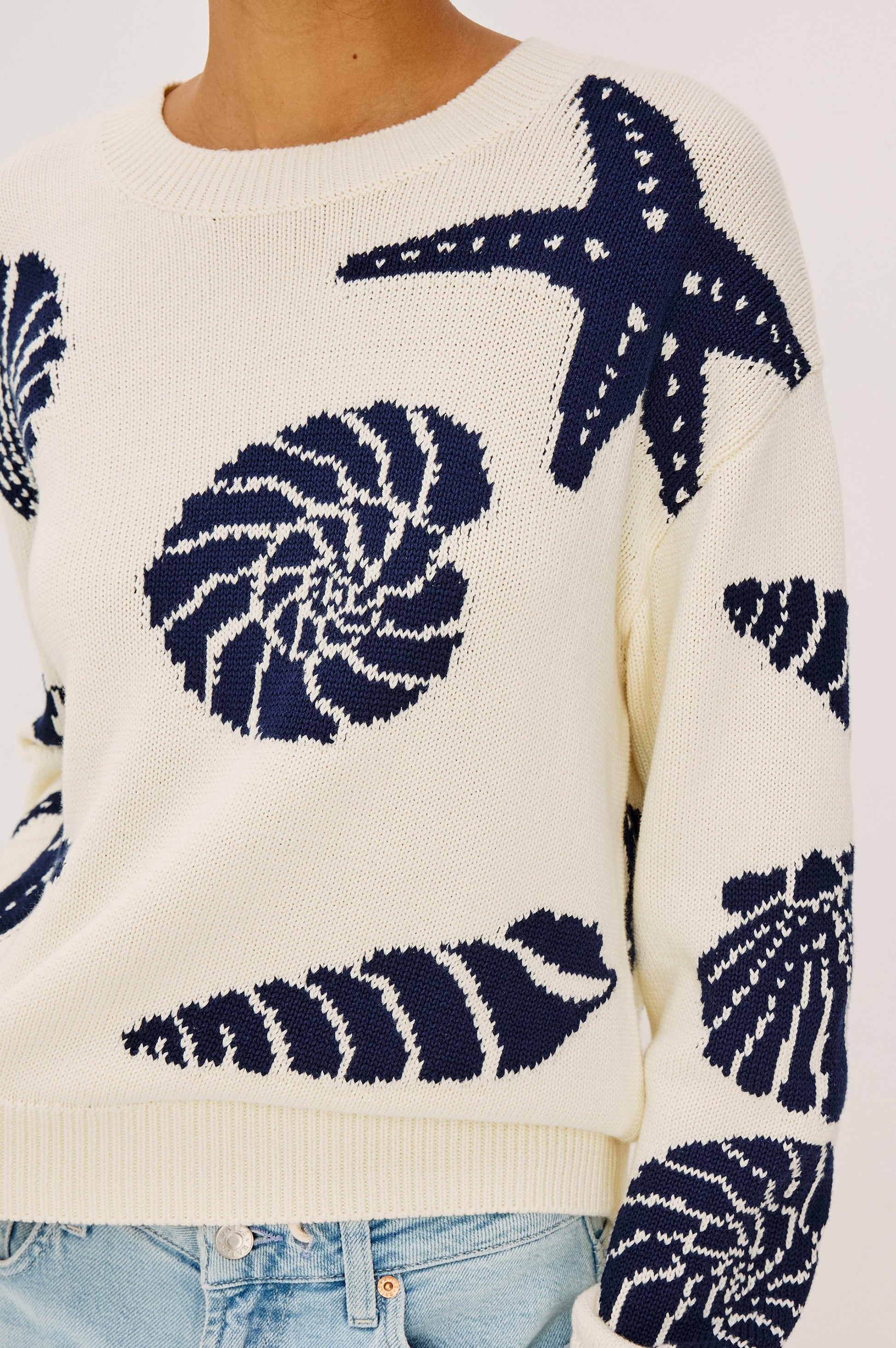A person wears the Zoey Shell Sweater by Rails, a cream crewneck embroidered with navy sea-themed designs like starfish and seashells, paired with light blue jeans.