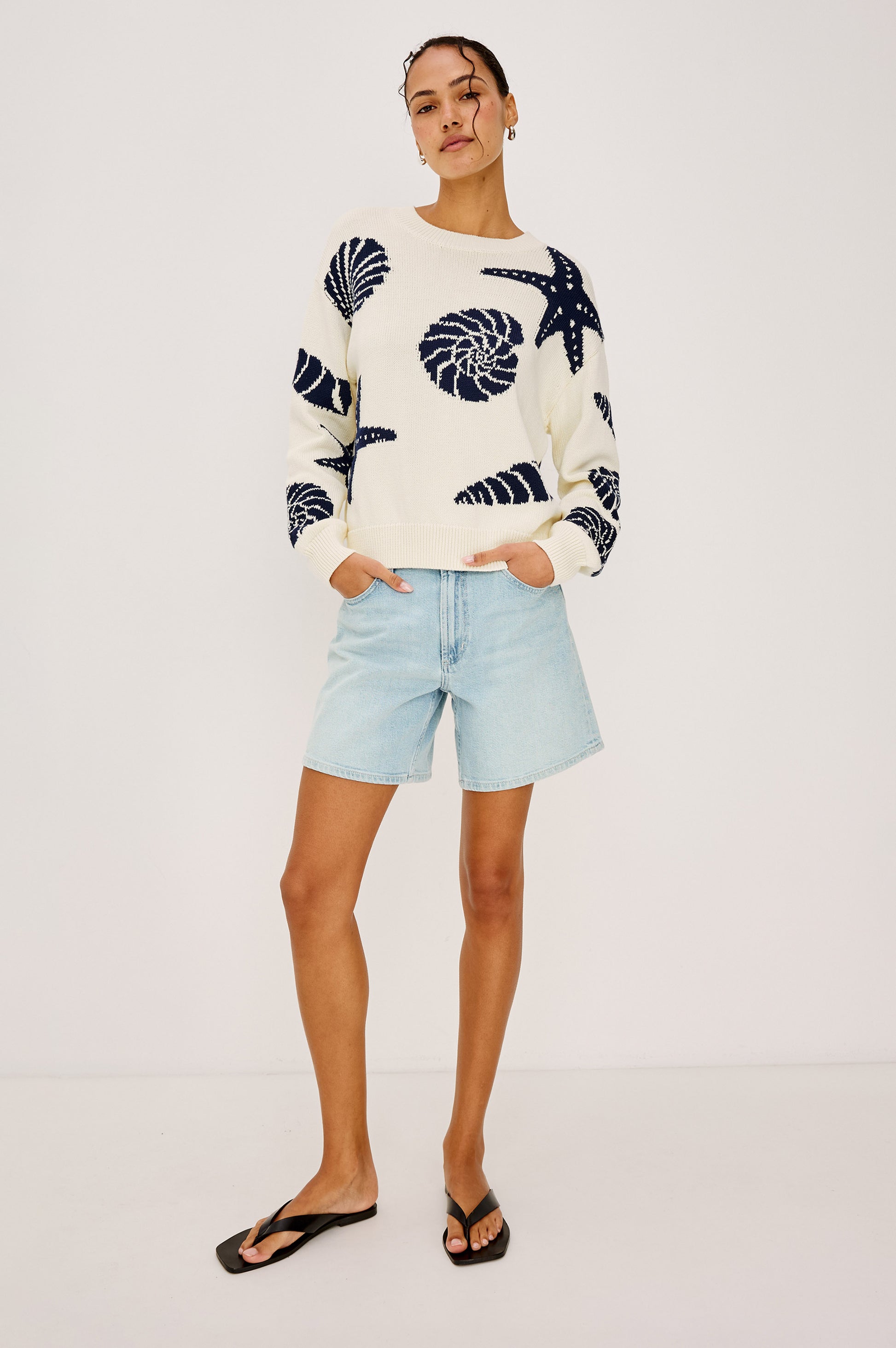 A person stands against a plain white background wearing the Rails Zoey Shell Sweater, featuring navy blue sea shell and starfish patterns, paired with light blue denim shorts and black flip-flops, hands in pockets and hair pulled back.