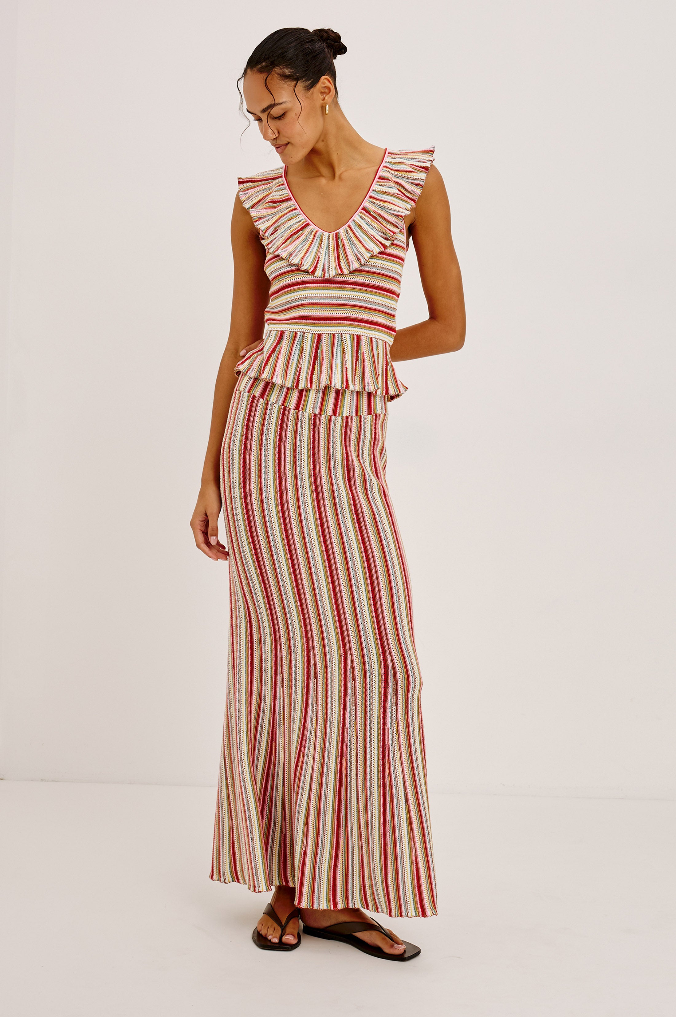A woman stands in a minimalist room wearing the Carmelita dress by Rails—a sleeveless, floor-length piece with a ruffled V-neckline and vertical red, white, and beige stripes. She smiles softly, hair tied back and sandals finishing her look.