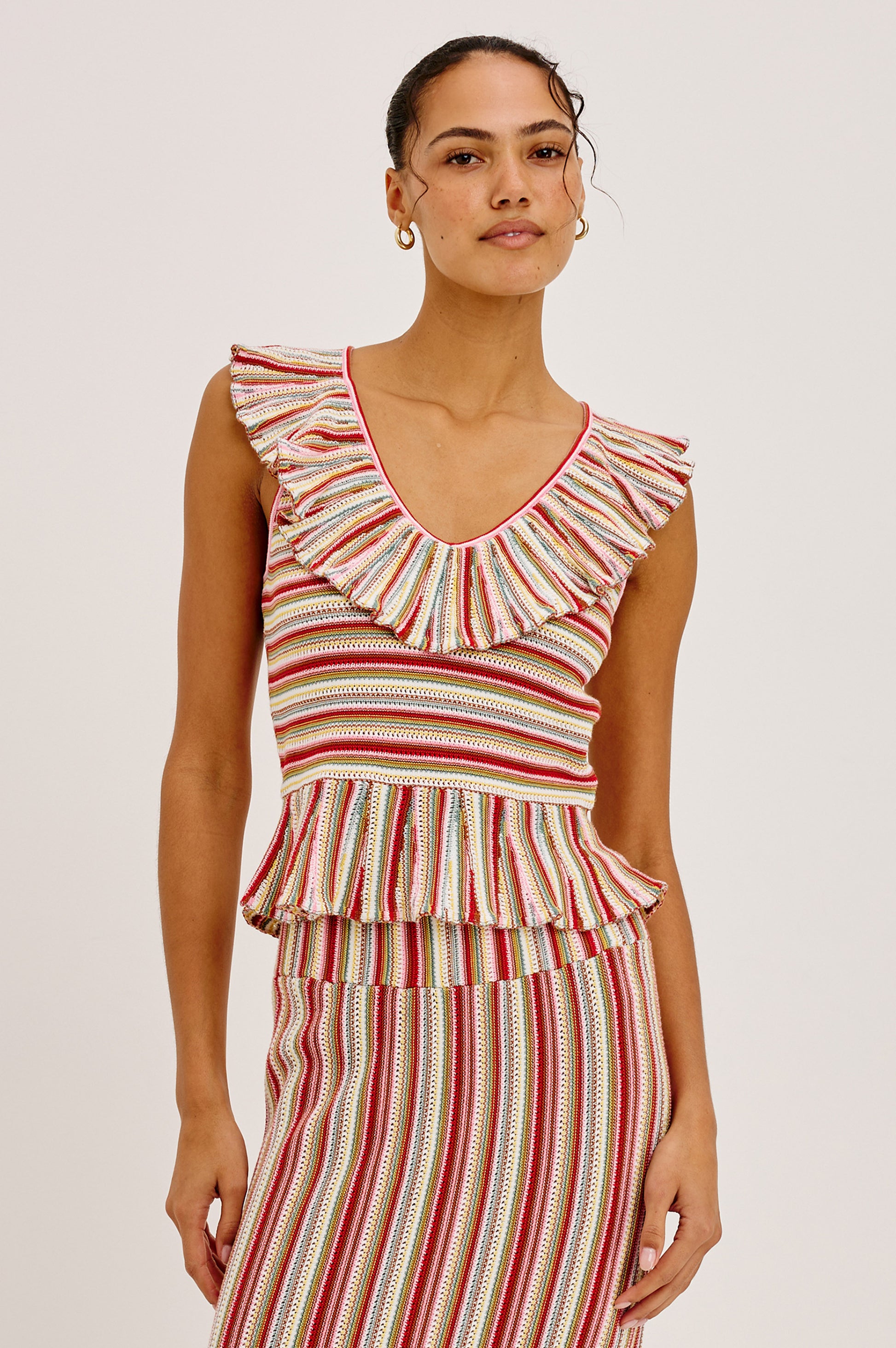 A woman wears the Rails Carmelita set in Red Multi—a sleeveless, striped crochet top with a ruffled V-neck and peplum hem, paired with a matching skirt. Her hair is tied back as she stands against a plain background.