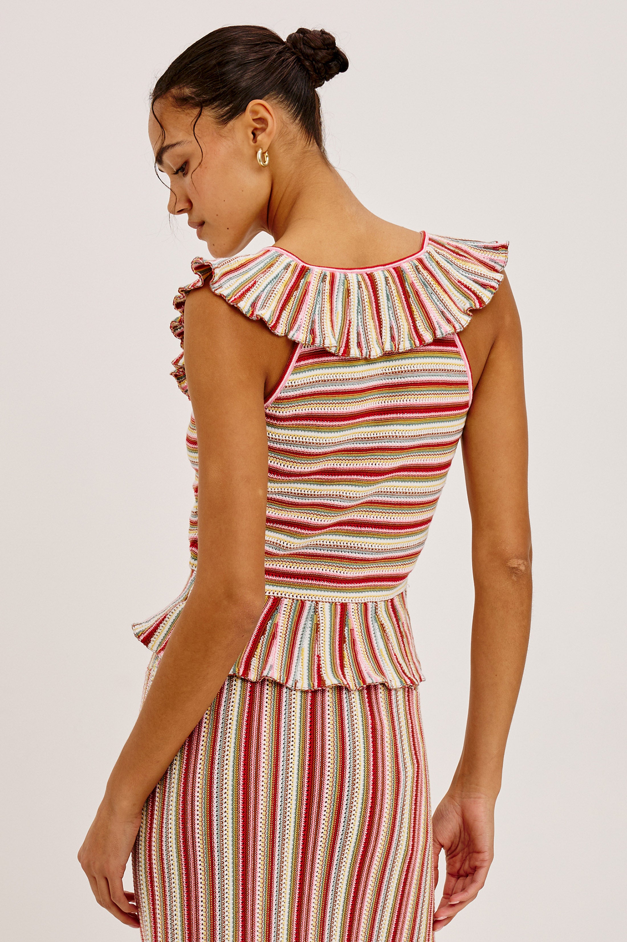 A woman with dark hair in a bun models the Rails Carmelita: a sleeveless, striped crochet top and skirt set featuring a ruffled collar and hem in red, white, and yellow tones. She stands with her back turned, glancing over her shoulder.