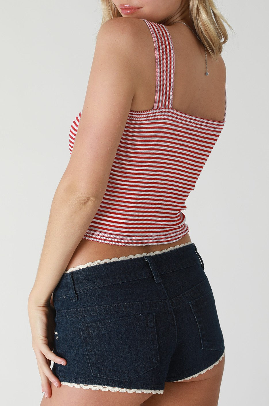 A woman stands sideways wearing the Niki Biki Modal Stripe Square Neck Crop, a red and white striped modal nylon spandex top, paired with dark denim shorts with lace trim. Her blonde hair is partially visible against a light background.
