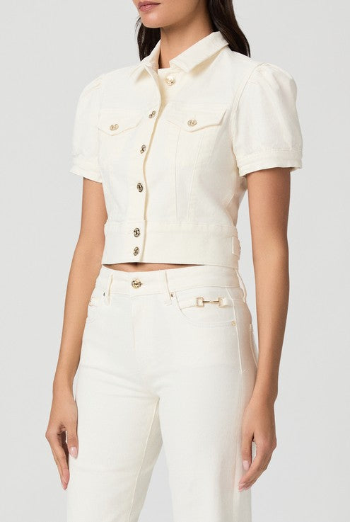 A woman models the Paige Kendra Denim Jacket in Ecru, an off-white, short-sleeve button-up style with double chest pockets and gold buttons, paired with matching high-waisted pants; set against a plain light gray background.