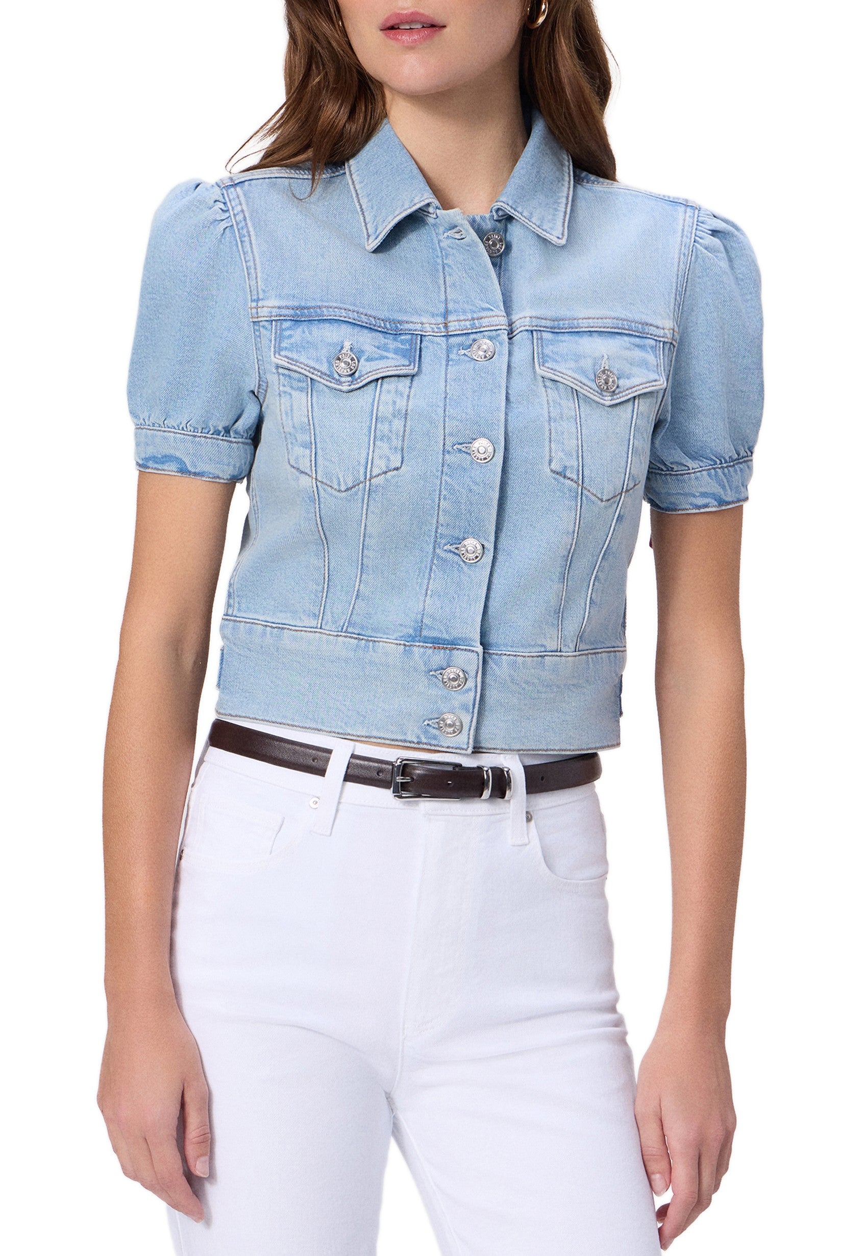 A woman wears the Paige Kendra Denim Jacket in Glide, a light blue, short-sleeve style with double chest pockets and front buttons, paired with high-waisted white pants and a dark belt. Her head is partially out of frame.