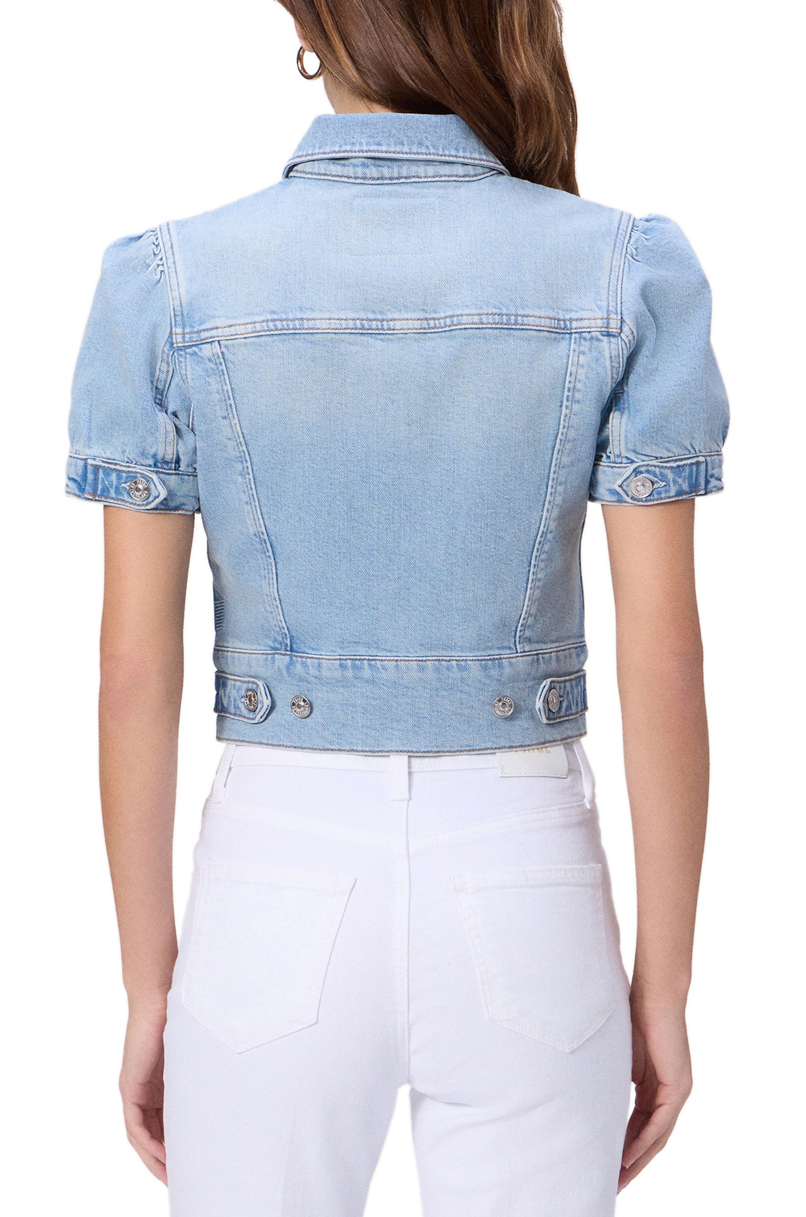 A woman with long, wavy brown hair is seen from the back wearing the Paige Kendra Denim Jacket in Glide—a light blue, heritage-inspired style featuring short puff sleeves—paired with white high-waisted jeans.