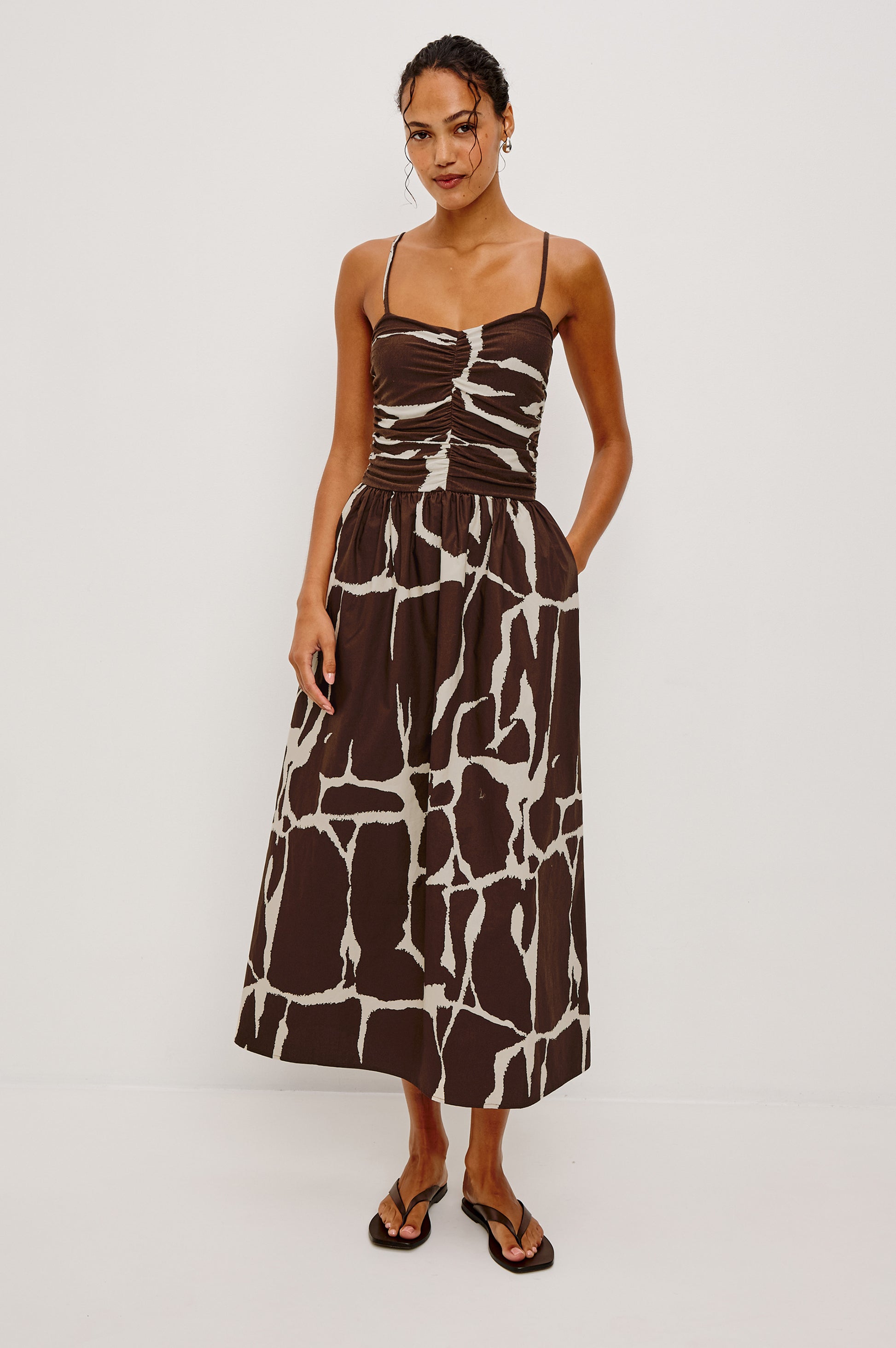 A woman smiles with her hands in the pockets of the Rails Fisher Midi Dress, featuring a brown and white giraffe print, spaghetti straps, a ruched bodice, and black flip-flops against a white background.