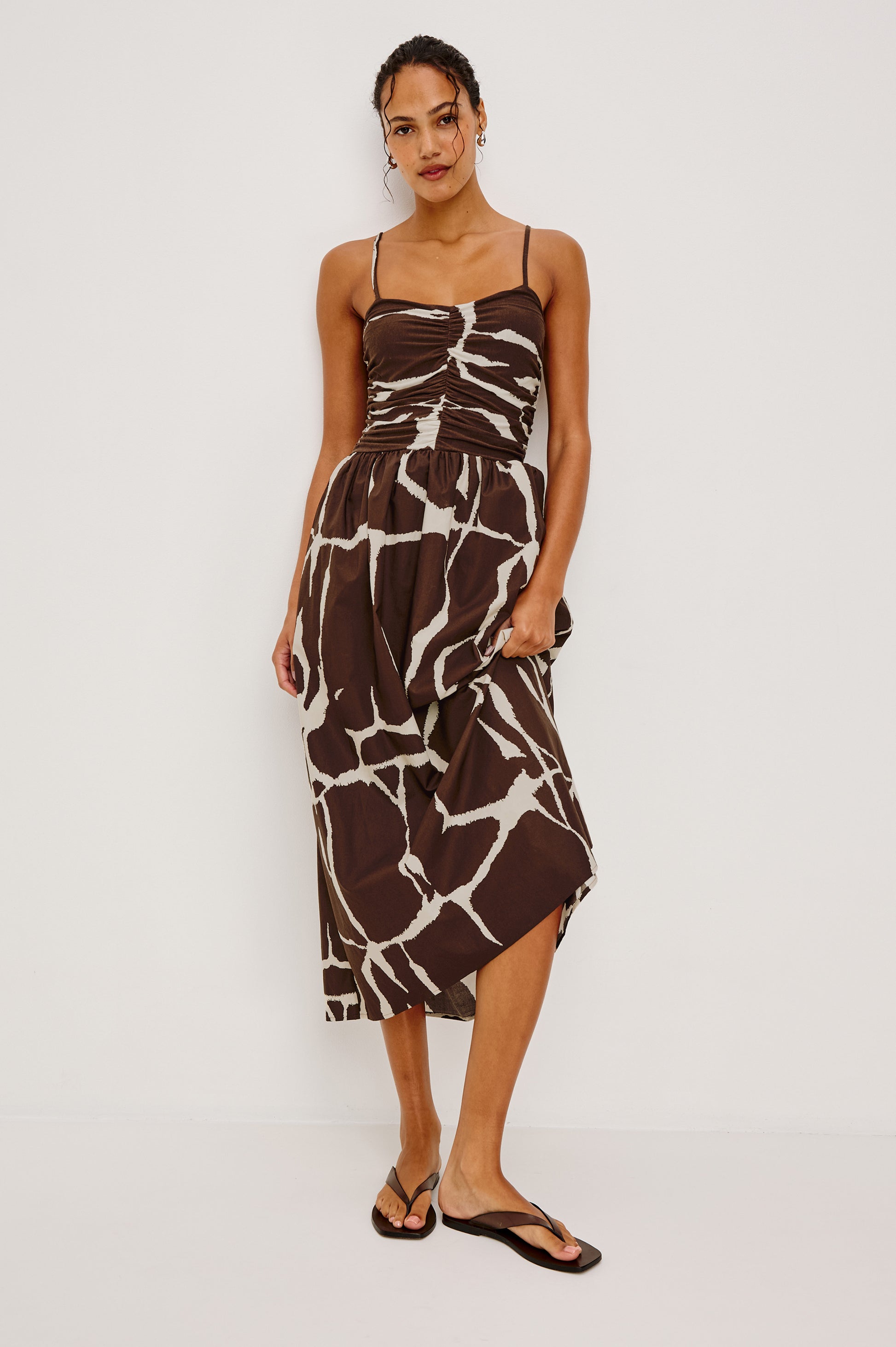 A woman wearing the Rails Fisher Midi Dress, featuring a brown and white abstract pattern, sleeveless design, ruched bodice, thin straps, and paired with black flip-flops stands against a plain white background.