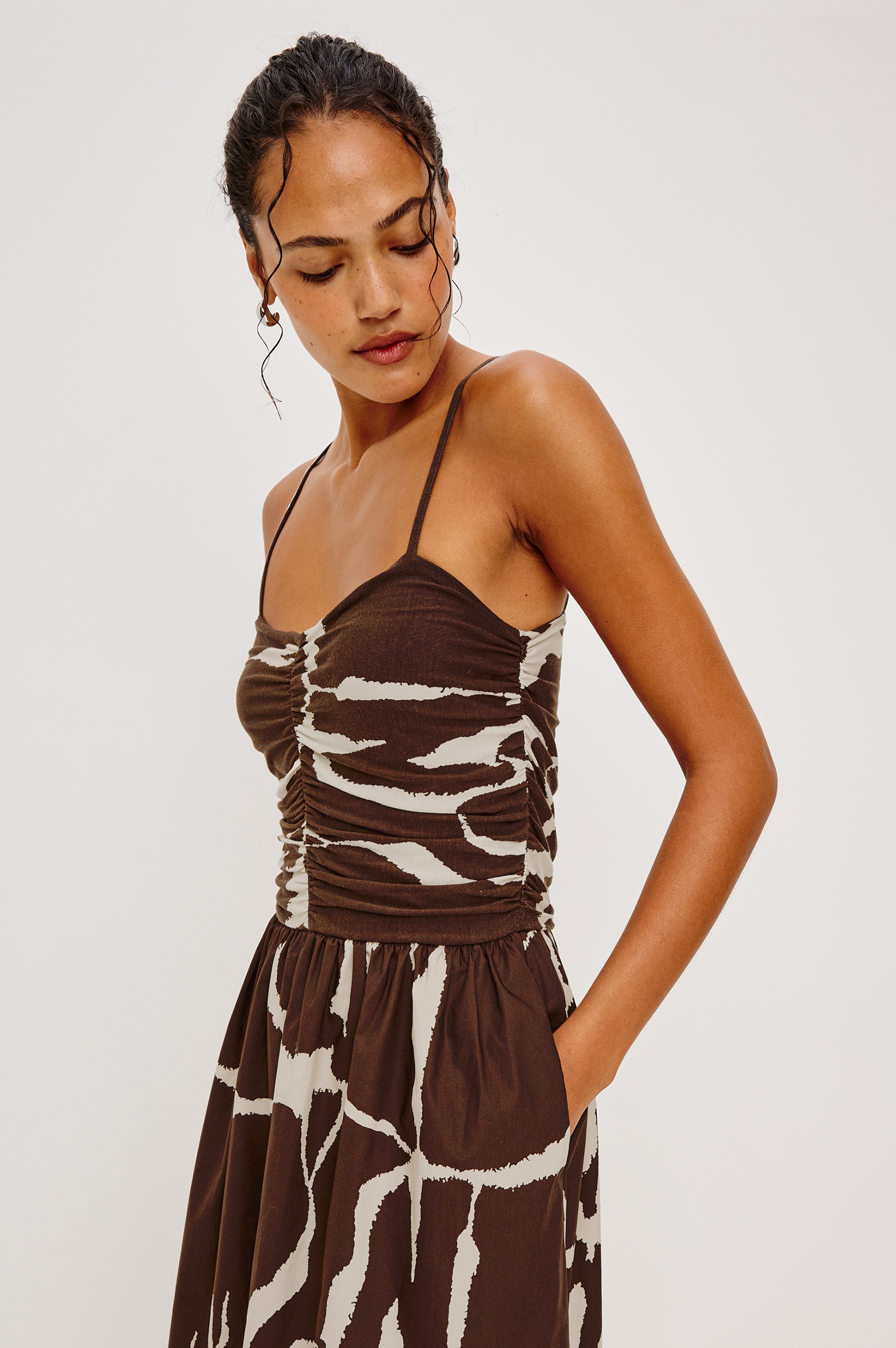 A woman wearing the Rails Fisher Midi Dress, featuring a brown and white abstract pattern and ruched bodice, stands against a plain light background, looking down.