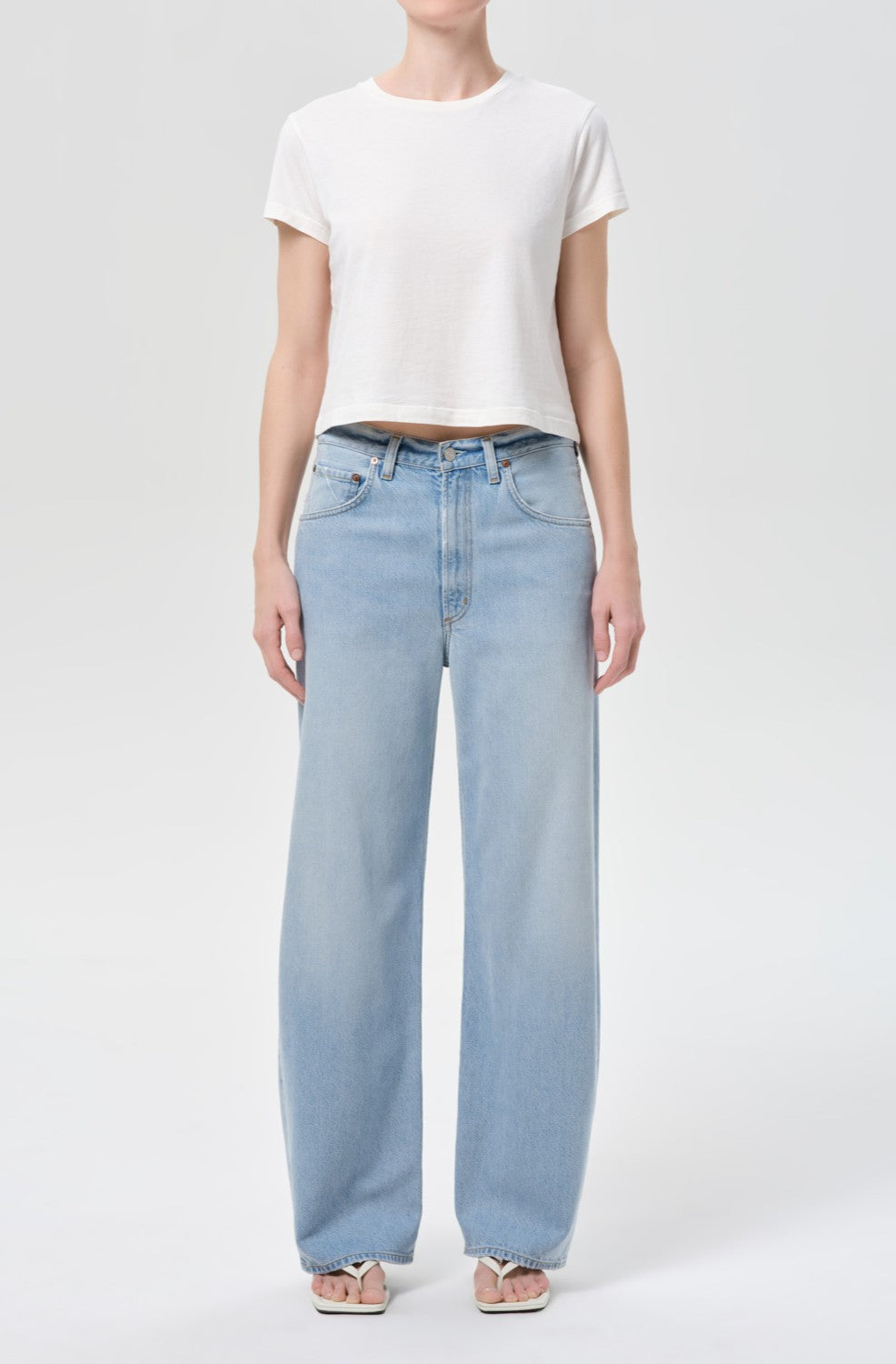 Wearing a plain white cotton crop top, the person pairs it with AGOLDE’s Low Curve Jean In Memory—a high-waisted, wide-leg, pale indigo denim pant—and open-toed sandals, standing against a light backdrop with their face not visible.