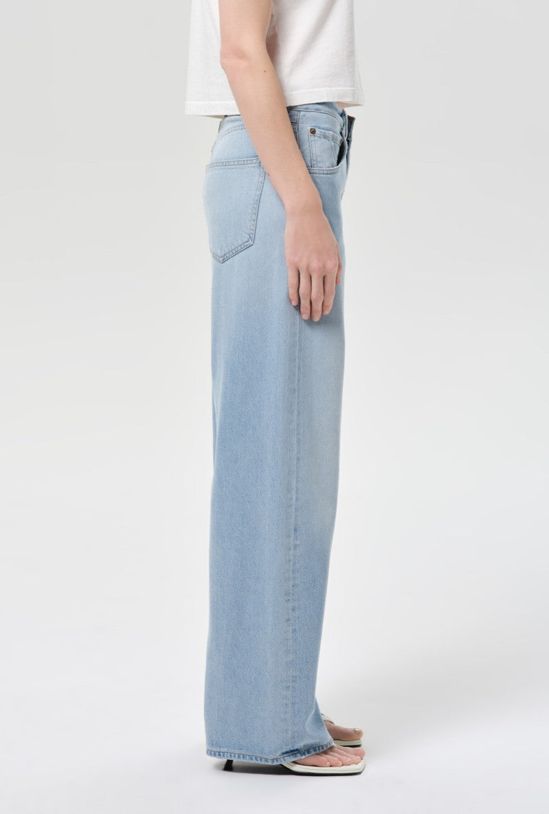 A person stands in profile against a plain light background wearing AGOLDE’s Low Curve Jean In Memory, made from pale indigo breathable cotton denim, paired with a white short-sleeve shirt and white open-toe sandals.