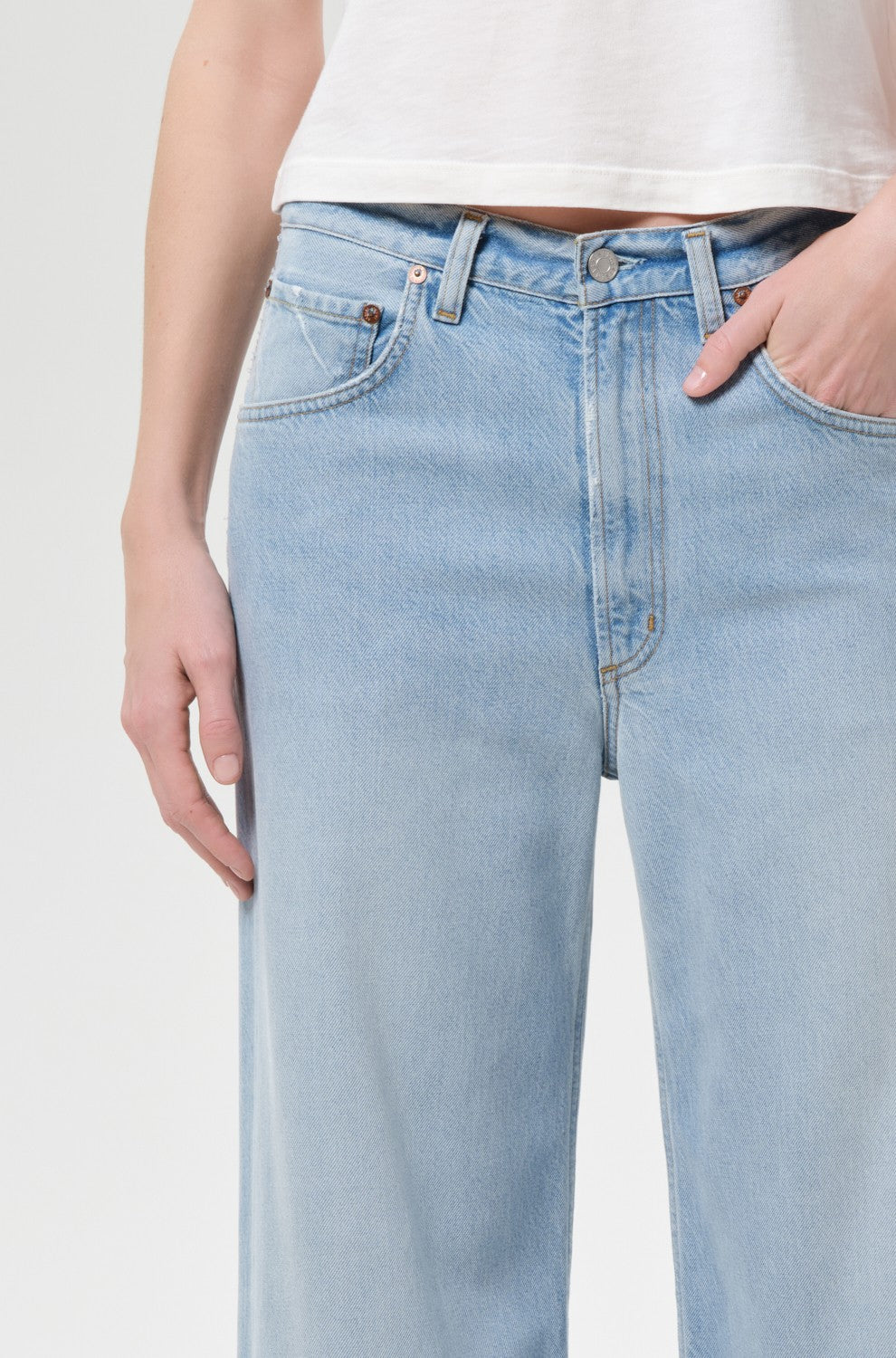 A person wearing AGOLDE's Low Curve Jean In Memory, made of breathable cotton in pale indigo, paired with a white crop top, stands with one hand in their pocket against a plain light background.