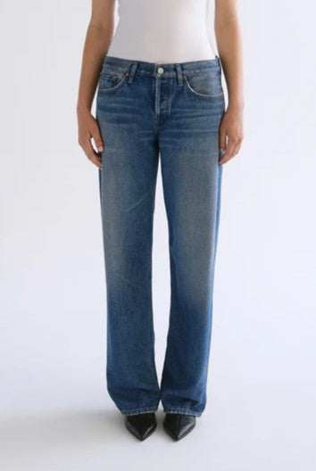 A person is wearing AGOLDE's Low Rise Loose In Expression jeans in the color EXPRESSION, crafted from ultra-soft denim with slight fading and a straight-leg fit. They are paired with a white sleeveless top and black pointed-toe shoes, standing against a plain light background.