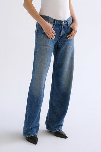 Wearing the AGOLDE Low Rise Loose In Expression jeans, characterized by their loose fit, lightweight denim, and slightly faded blue wash, a person stands with hands in pockets. The jeans feature a low-rise waist for a relaxed look and are paired with a white top and black pointed-toe shoes.
