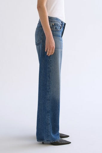 A person stands sideways against a plain white background, wearing the AGOLDE Low Rise Loose In Expression indigo denim pants styled with a white top and black pointed-toe shoes.