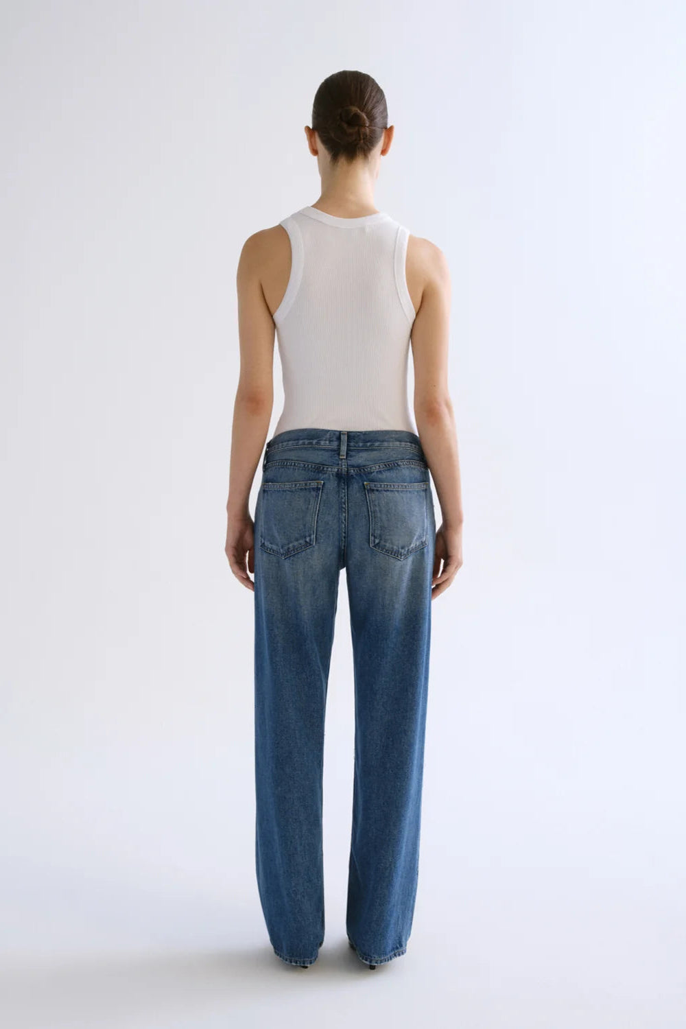 Person wearing white sleeveless tank top and low-rise medium-wash blue jeans, shown from back view.