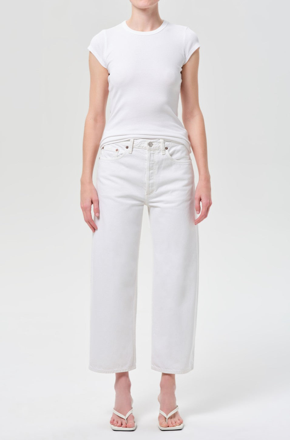 A person wearing a fitted white short-sleeve t-shirt, AGOLDE Arc Jean 29 Inseam in Chalk White, and white heeled sandals stands against a plain light background.