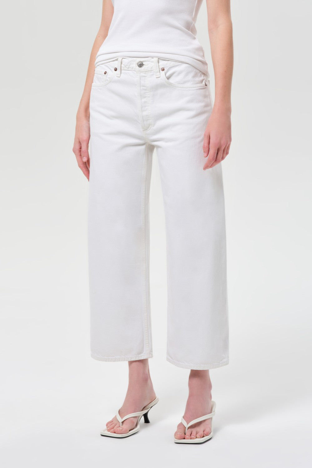 A person wearing a white sleeveless top, AGOLDE Arc Jean 29 Inseam in Chalk White, and white heeled sandals stands against a plain light background. The person's head is not visible in the image.