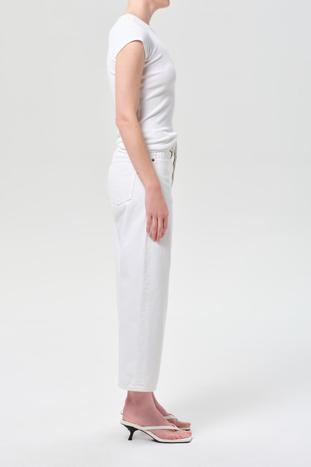 A person stands in profile against a plain background, wearing the AGOLDE Arc Jean 29 Inseam in Chalk White, a white short-sleeve top, and white heeled sandals. Their arms are at their sides and their face is not visible.