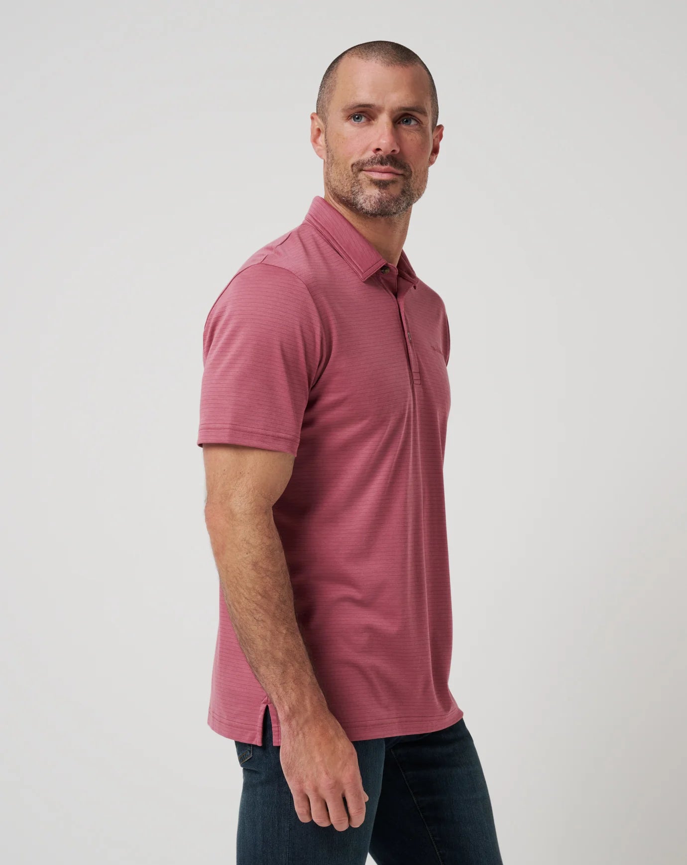 A man with short hair and a beard wears The Zinna 2.0 rose polo shirt by TravisMathew and dark jeans, standing against a plain light background, looking slightly to the side.