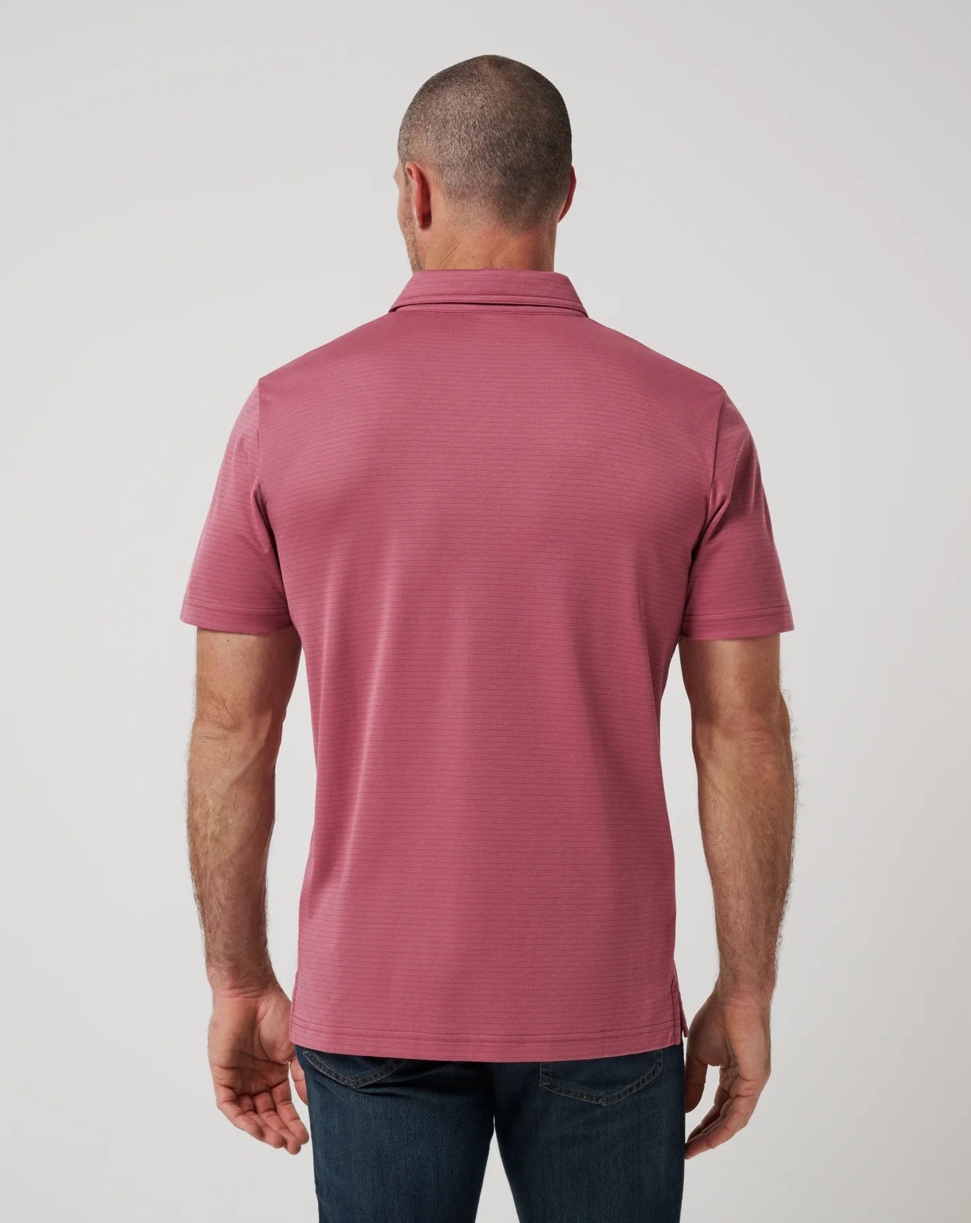 A man stands with his back to the camera wearing TravisMathew’s The Zinna 2.0, a short-sleeved rose polo shirt known for lightweight breathability, paired with dark blue jeans against a plain light background.