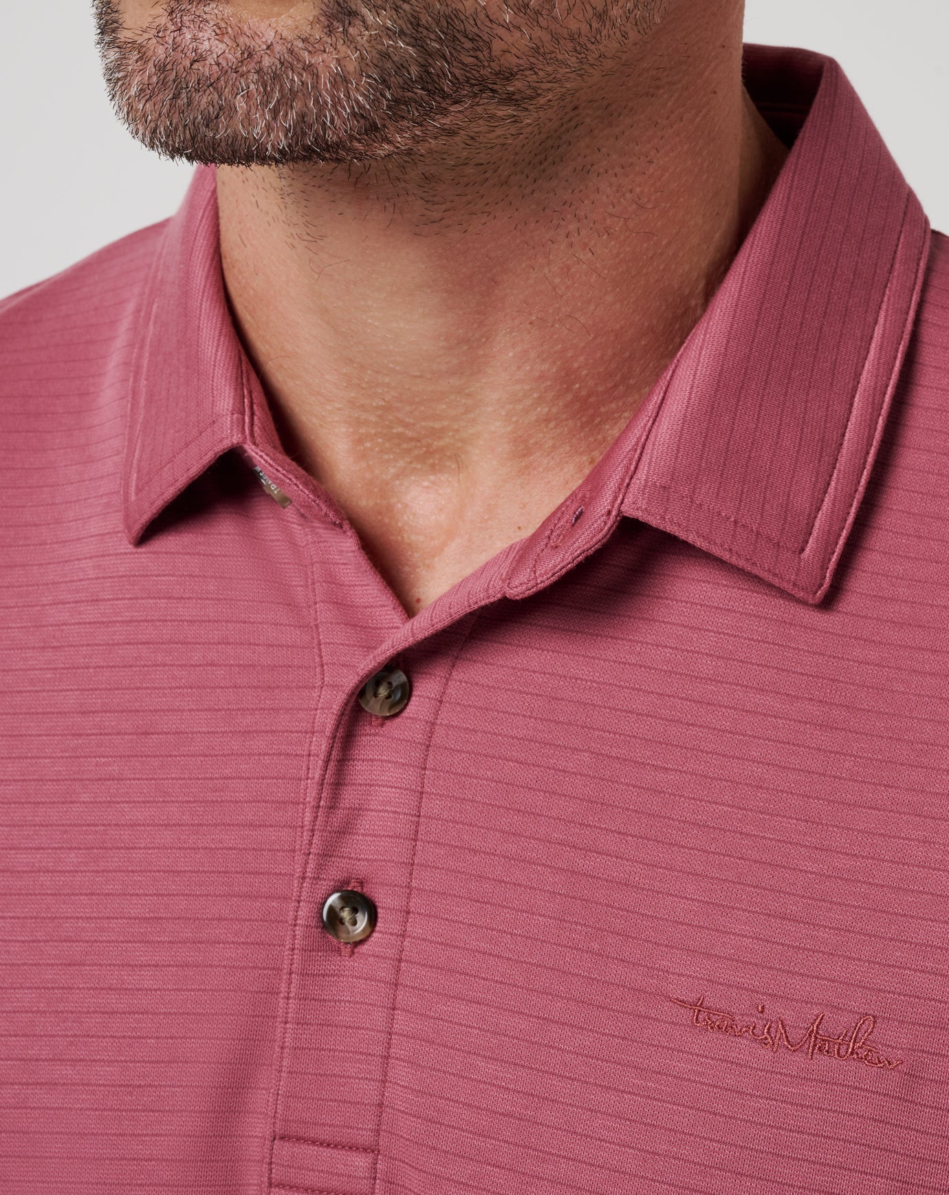 Close-up of a man in The Zinna 2.0 by TravisMathew—a ribbed, mauve polo shirt with lightweight breathability, button placket, and a subtle embroidered “TravisMathew” logo on the chest. His trimmed beard and mustache are partially visible.