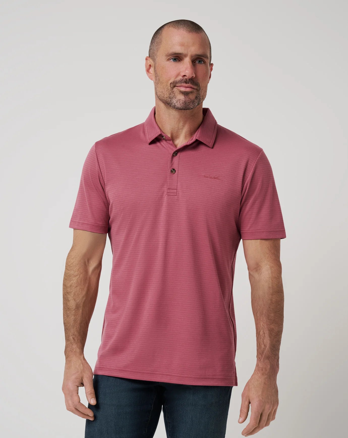 A man with short hair and a trimmed beard wears the TravisMathew Zinna 2.0 polo in red with a double-needle collar and dark jeans. He stands against a plain light background, looking slightly to the side.