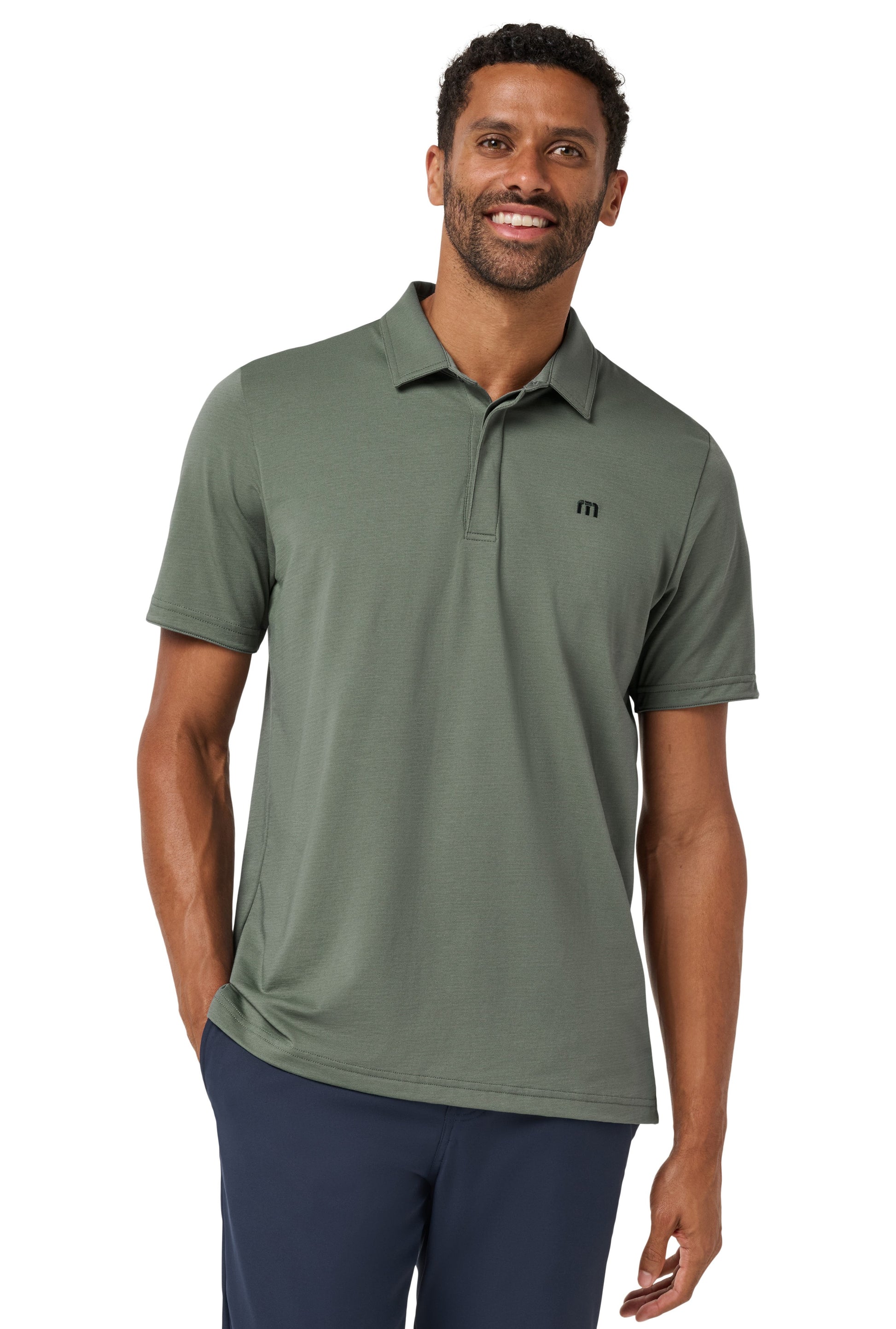 A man with short curly hair and a beard smiles at the camera, wearing navy pants and the TravisMathew Heater Series Jersey Polo in BALSALM GREEN performance fabric, featuring a small black chest logo, with one hand in his pocket.