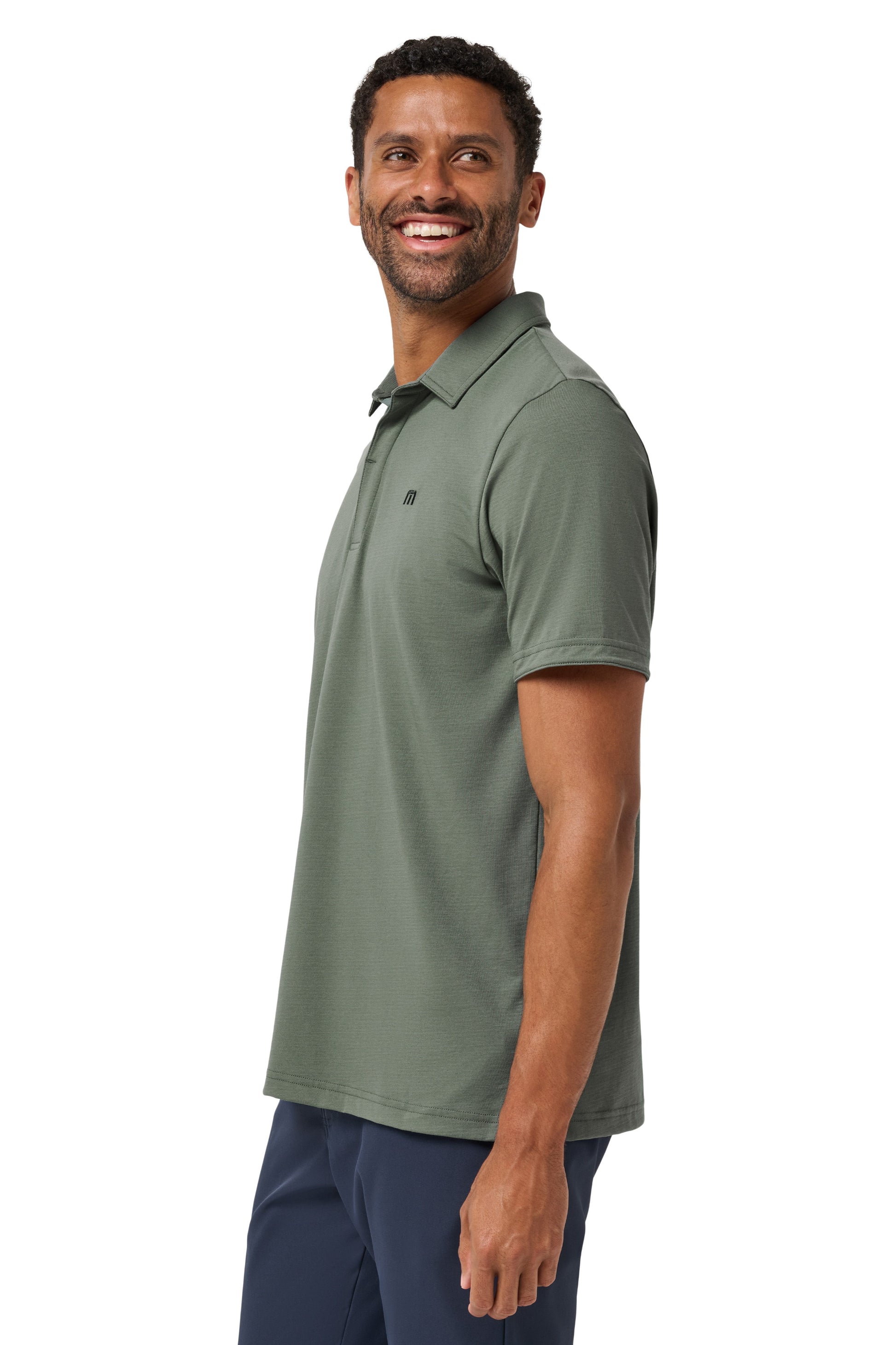 A man with short dark hair and a beard smiles in a relaxed pose against a white background, wearing navy pants and the TravisMathew Heater Series Jersey Polo in sage green.