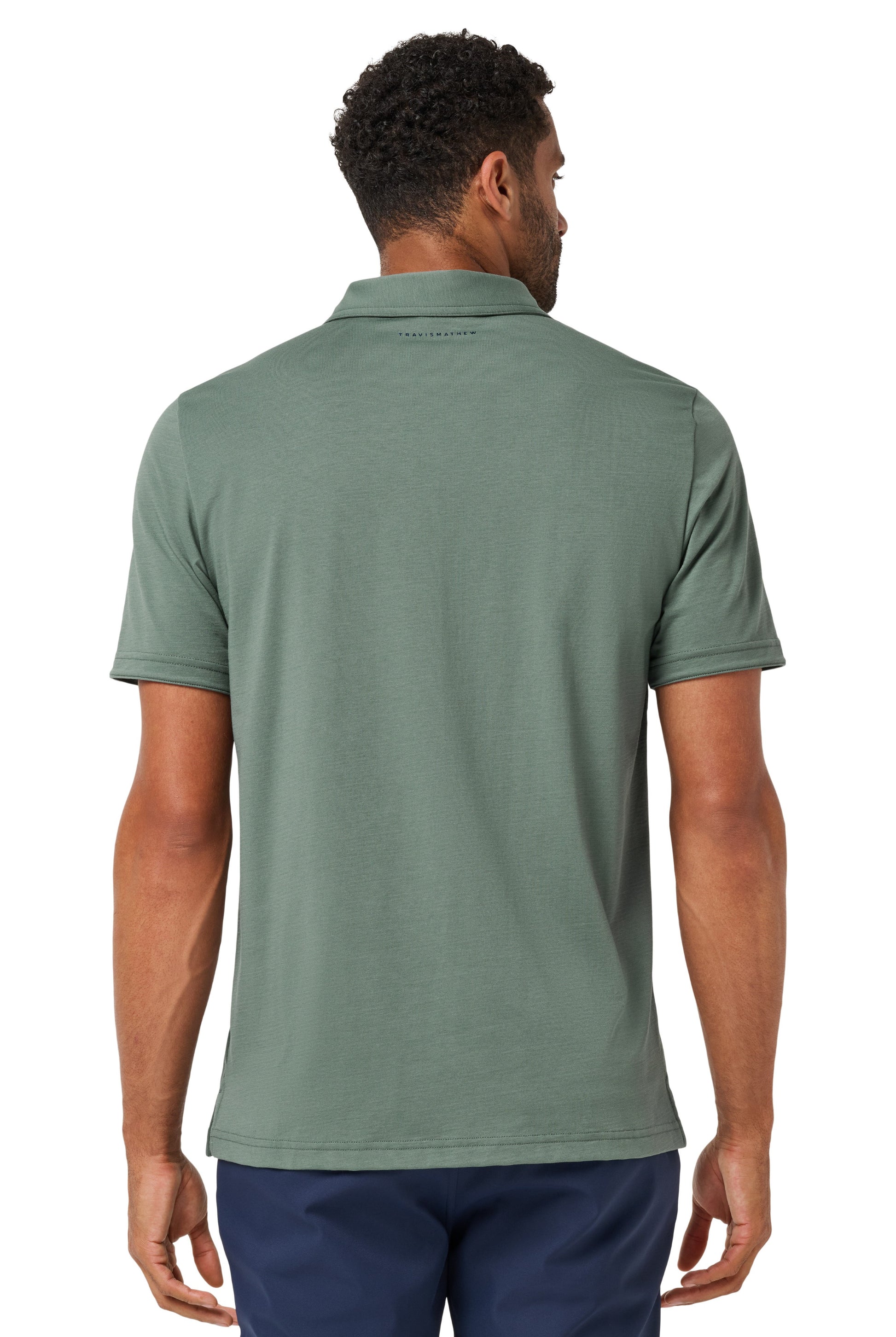A man is seen from behind wearing a TravisMathew Heater Series Jersey Polo in sage green with navy blue pants, both made of breathable performance fabric. The background is plain white.