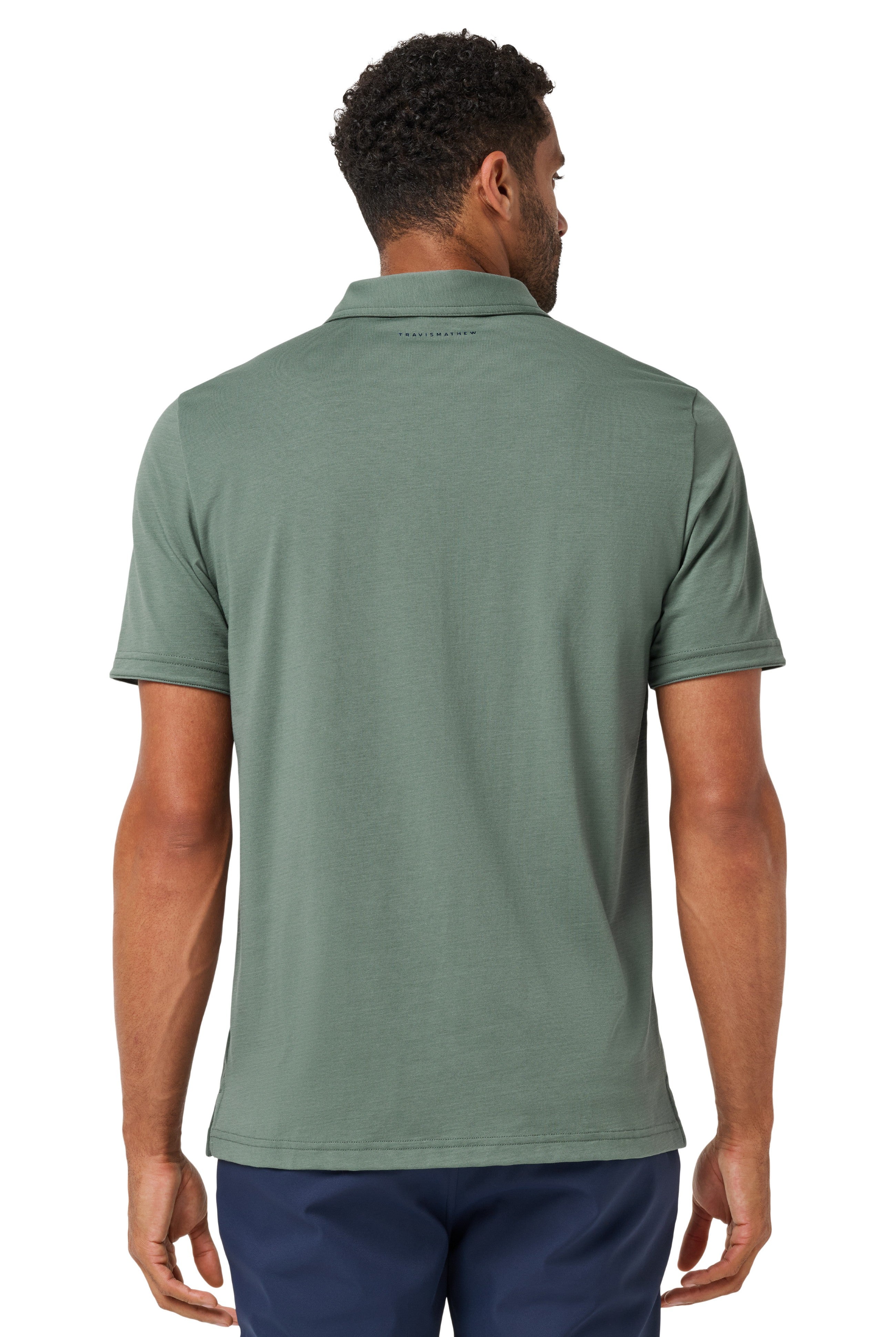 A man is seen from behind wearing a TravisMathew Heater Series Jersey Polo in sage green with navy blue pants, both made of breathable performance fabric. The background is plain white.