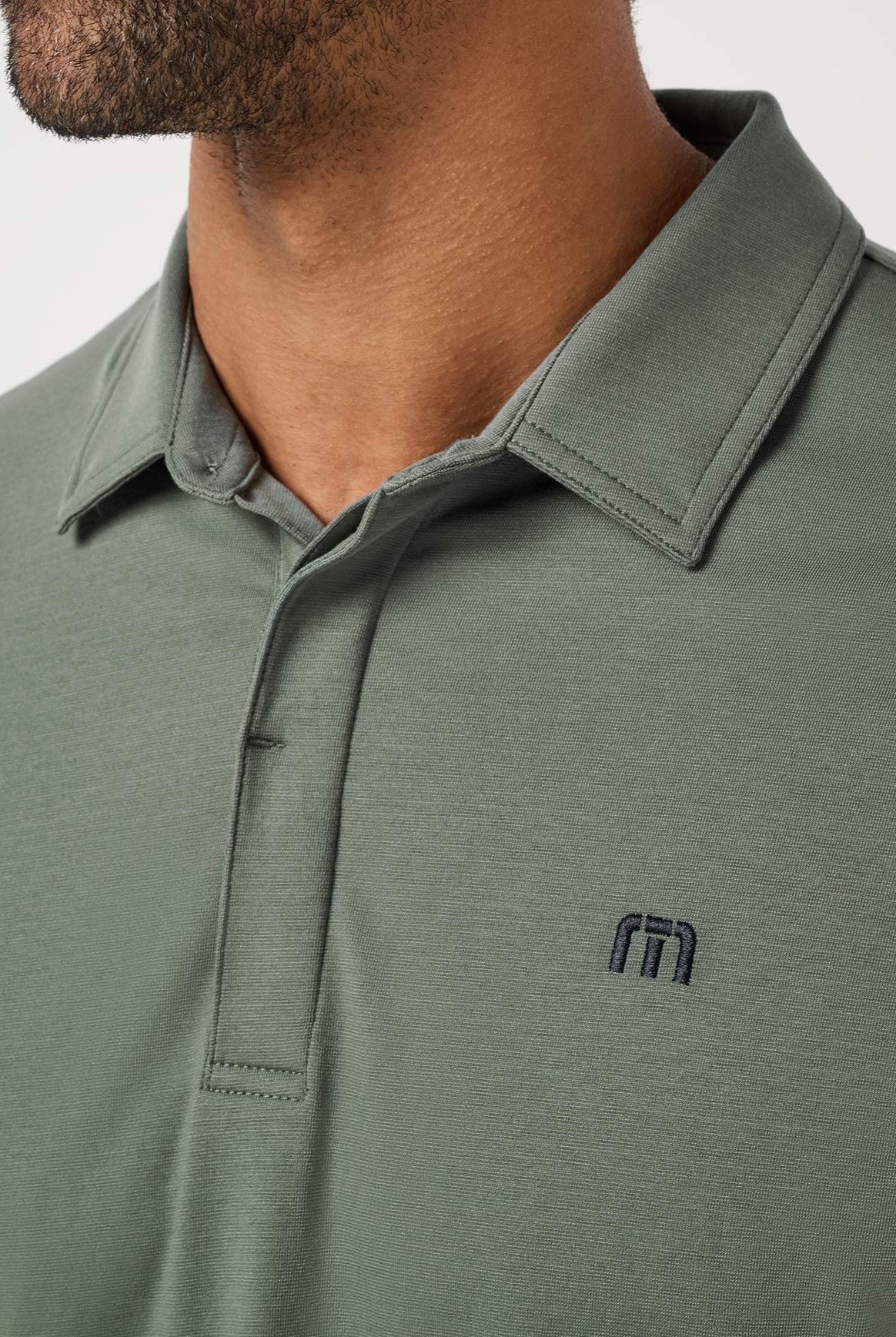Close-up of a person in the TravisMathew Heater Series Jersey Polo, a sage green, collared golf shirt made from breathable performance fabric with a small dark embroidered chest logo; only lower face, neck, and upper torso are shown.