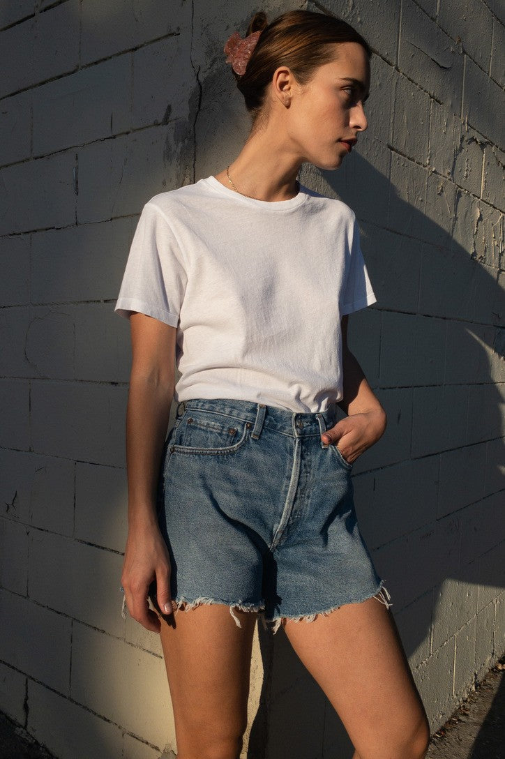 A person stands against a sunlit, textured wall, wearing a white t-shirt tucked into AGOLDE's Parker Long Denim Short in Skywave—vintage-inspired, non-stretch high-waisted shorts—with one hand in their pocket and hair pulled back with a scrunchie.