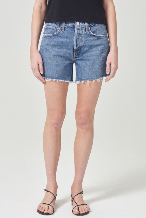 Someone stands against a plain light background, wearing AGOLDE’s Parker Long Denim Short in Skywave with a black top and black strappy sandals. The photo, cropped from the shoulders down, highlights the non-stretch cotton shorts.
