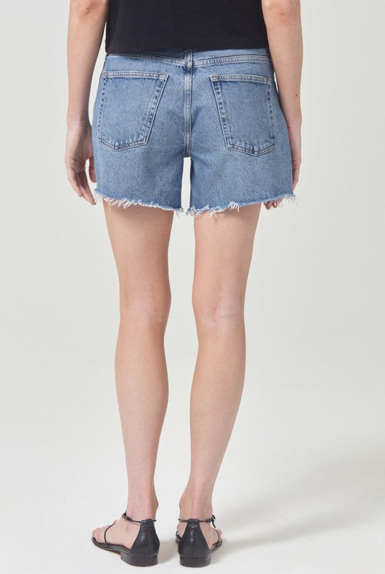 Person wearing a black top, AGOLDE Parker Long Denim Short in Skywave featuring frayed hems and non-stretch cotton, with black sandals, shown from the back against a plain light background.