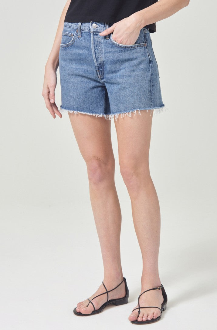 A person stands against a plain white background wearing a black sleeveless top, AGOLDE Parker Long Denim Short in Skywave, and black strappy sandals.