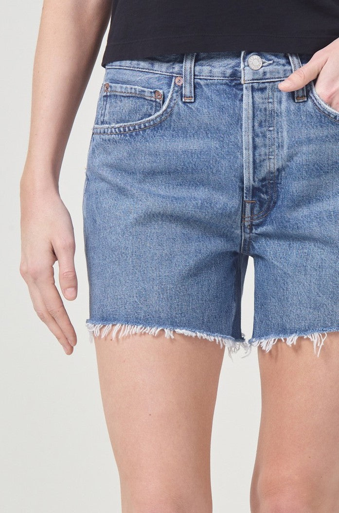 A person in a black top and AGOLDE Parker Long Denim Short In Skywave, crafted from non-stretch cotton with frayed hems, stands with one hand partially in a pocket.