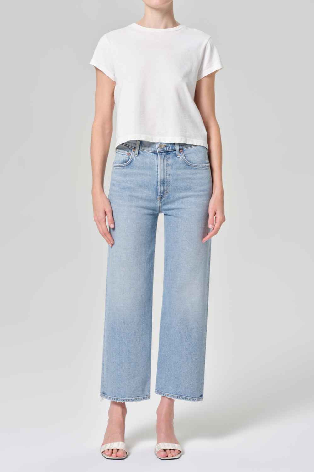 A person stands against a plain background, wearing AGOLDE’s Harper Crop In Formula—a white short-sleeve cropped t-shirt—paired with high-waisted light blue wide-leg jeans and white open-toe sandals.