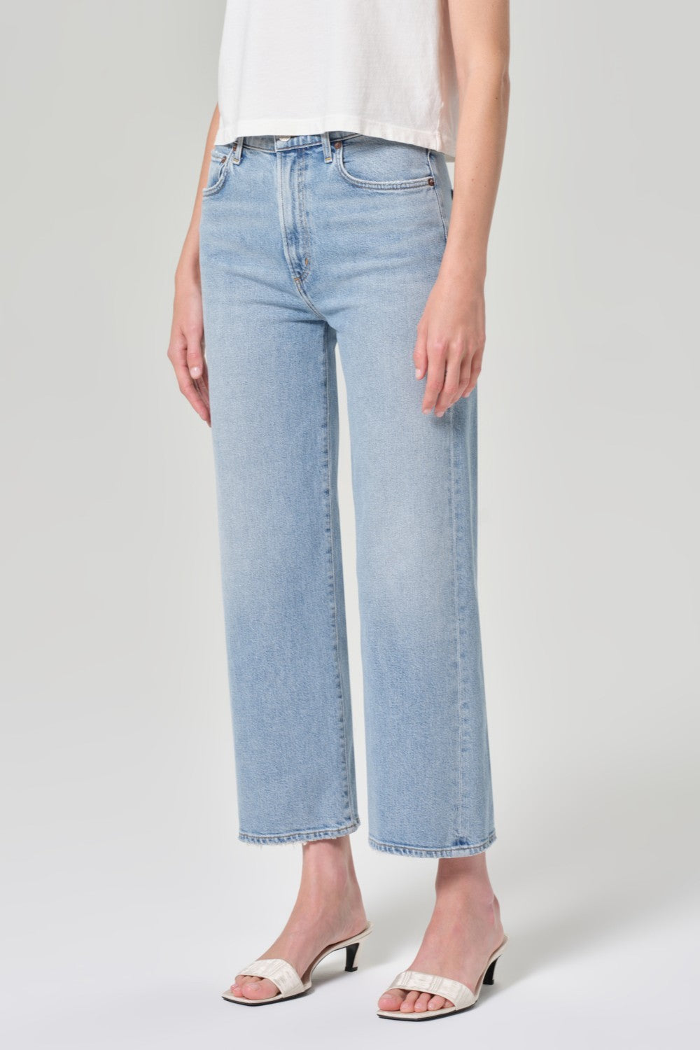 A person stands against a plain light background wearing AGOLDE's Harper Crop In Formula jeans in light denim, paired with a white sleeveless top and white heeled sandals.