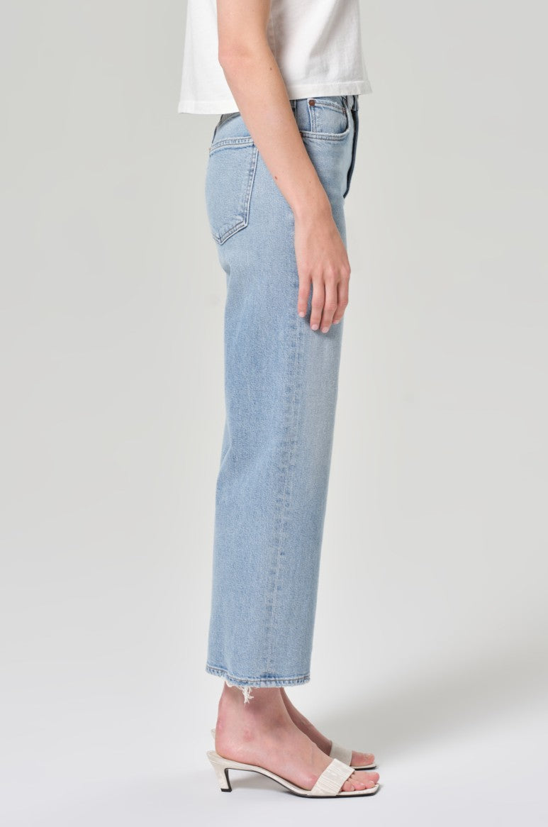 A person stands sideways against a light gray background wearing AGOLDE's Harper Crop In Formula high-waisted light blue jeans, a white short-sleeve top, and white open-toe low-heeled sandals.