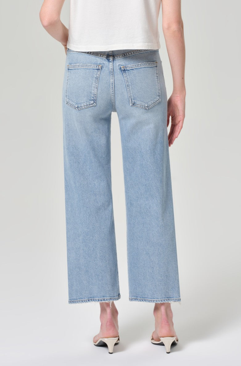A person wearing AGOLDE's Harper Crop In Formula, light blue straight-leg jeans, and white heeled sandals stands with their back to the camera. Only the lower half of their body is visible against a plain background.