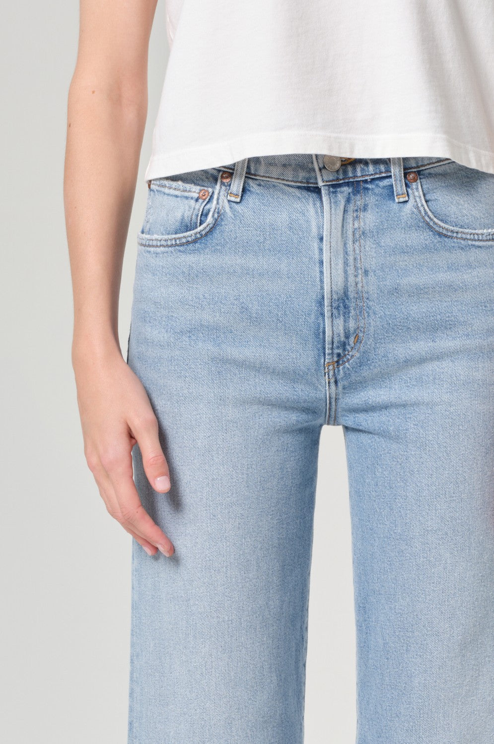 The AGOLDE Harper Crop In Formula features light blue, high-waisted comfort stretch denim jeans styled with a white sleeveless cropped top; the model stands from mid-torso to mid-thigh against a neutral background.
