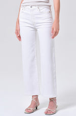 A person wears AGOLDE's Harper Crop In White Wash jeans—high-waisted, wide-leg denim made from regenerative cotton—paired with a light top and white strappy sandals, standing against a plain background. The photo is cropped at the shoulders.