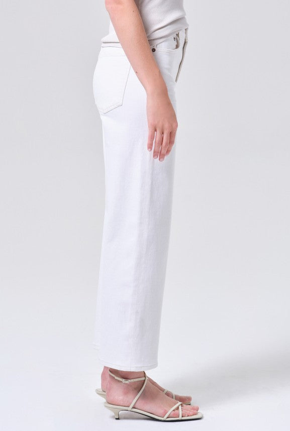 A person stands sideways in AGOLDE's Harper Crop In White Wash—white denim crafted from regenerative cotton—paired with a light ribbed tank and white thin-strap sandals. The background is plain and light-colored.