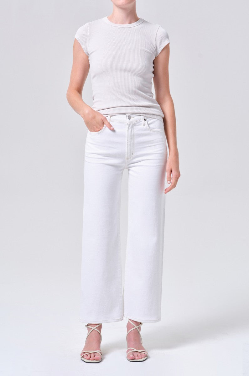 A person stands against a plain background, wearing a fitted white short-sleeve top, AGOLDE Harper Crop In White Wash jeans, and white strappy heeled sandals. One hand rests casually in their pocket.