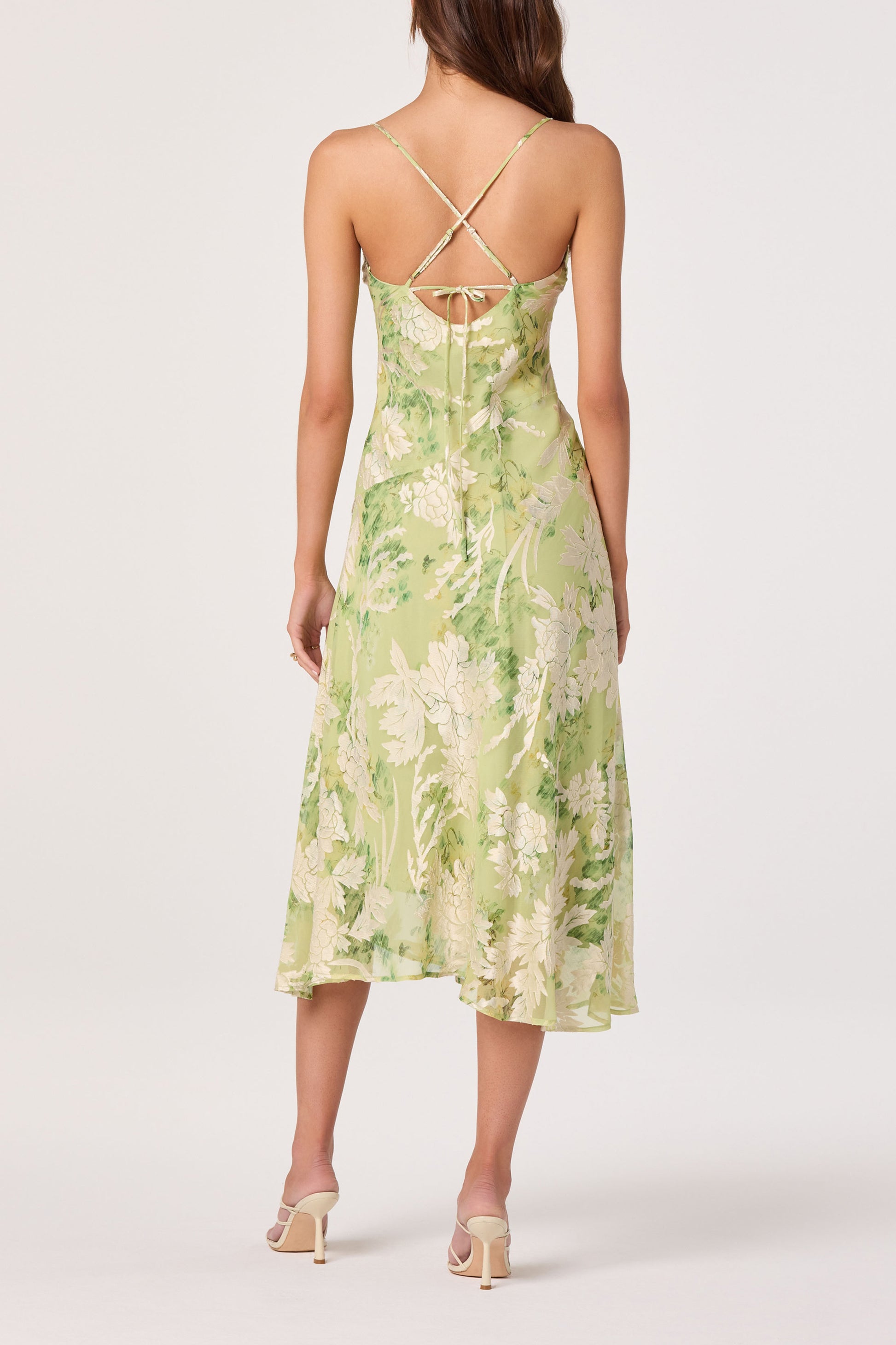 A woman stands facing away in the ASTR Gaia Dress—a light green, floral midi with thin crisscross back straps and a tie detail—paired with light-heeled sandals for an elegant look.