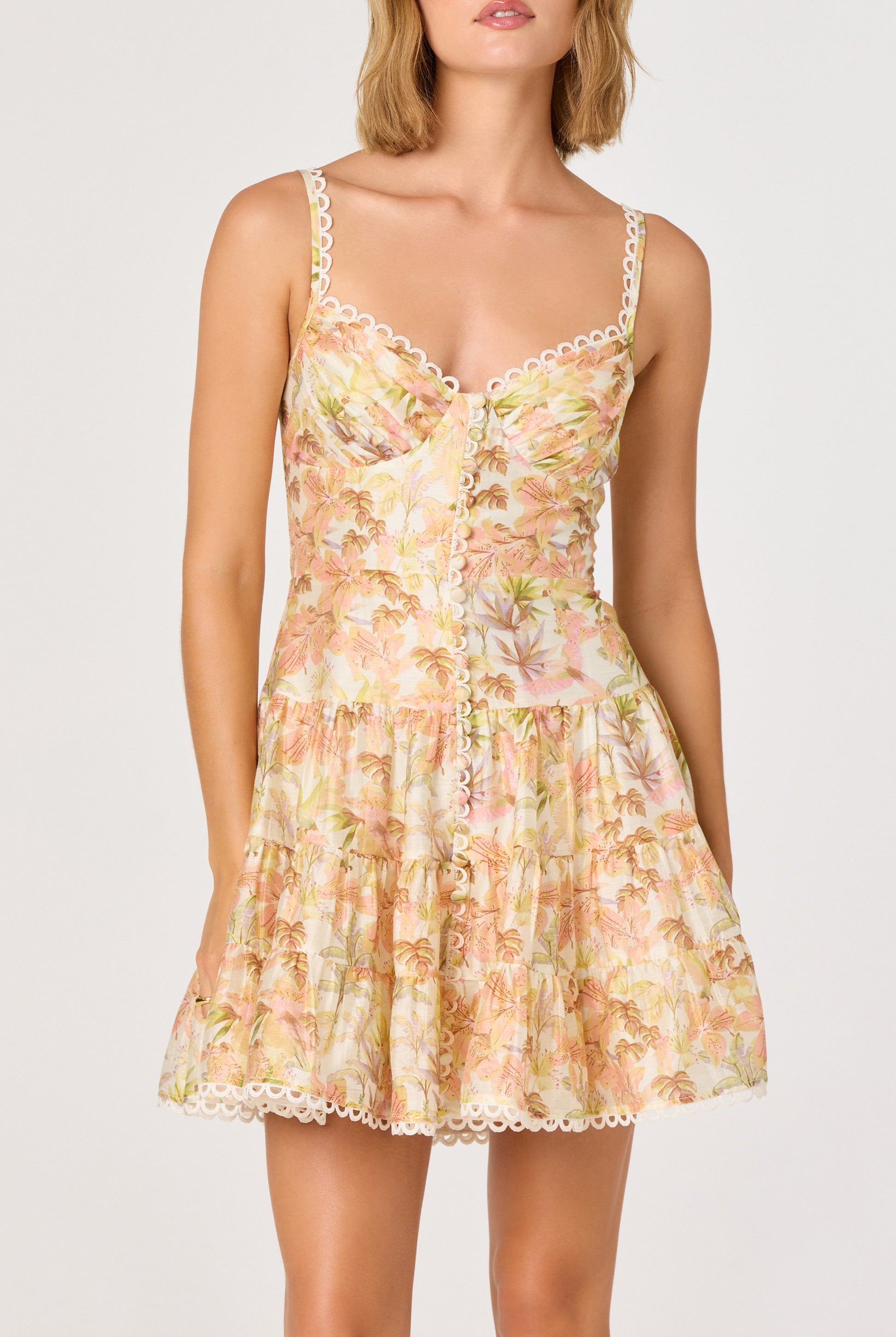 A woman wears the ASTR Tazia Mini Dress—sleeveless, floral print with lace trim, front buttons, fitted boned bodice, and a flared short skirt—standing against a plain light background.