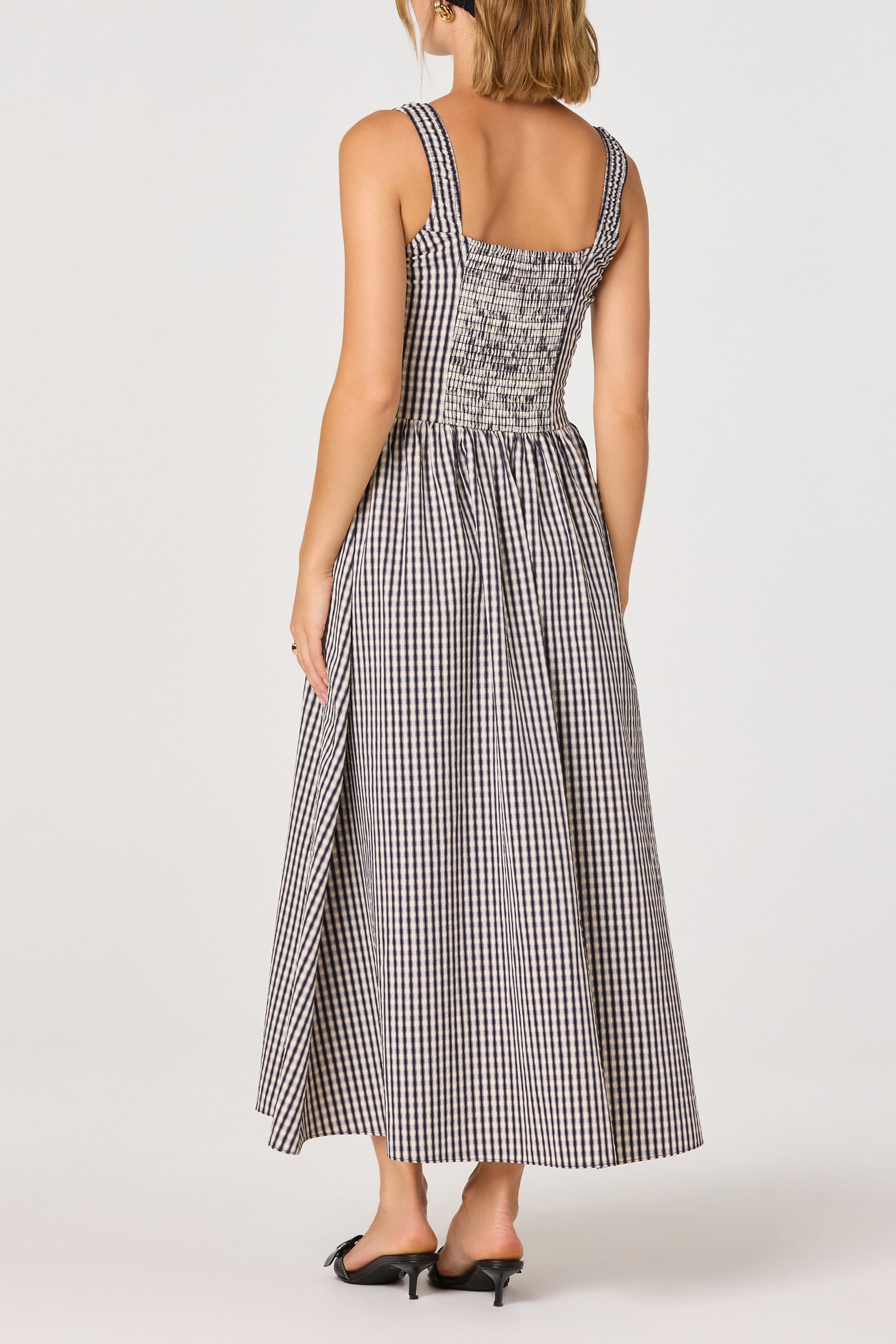 A woman is shown from the back on a plain background wearing the Temperance Maxi Dress by ASTR—a sleeveless, ankle-length, black-and-white checkered woven maxi dress with smocked tank straps—paired with black heels.